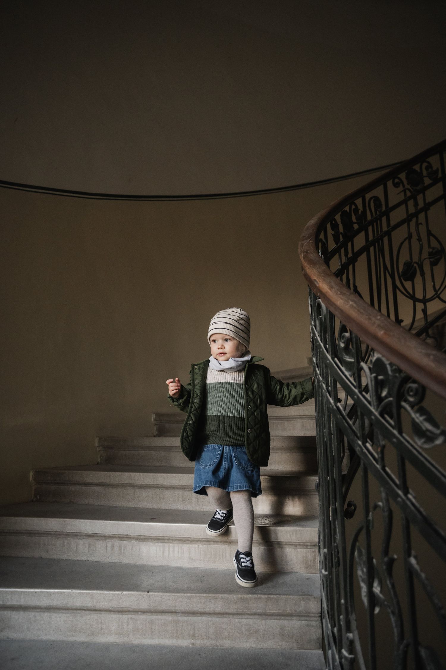 A small child in a winter coat sits on a dimly lit staircase with ornate metal railings.