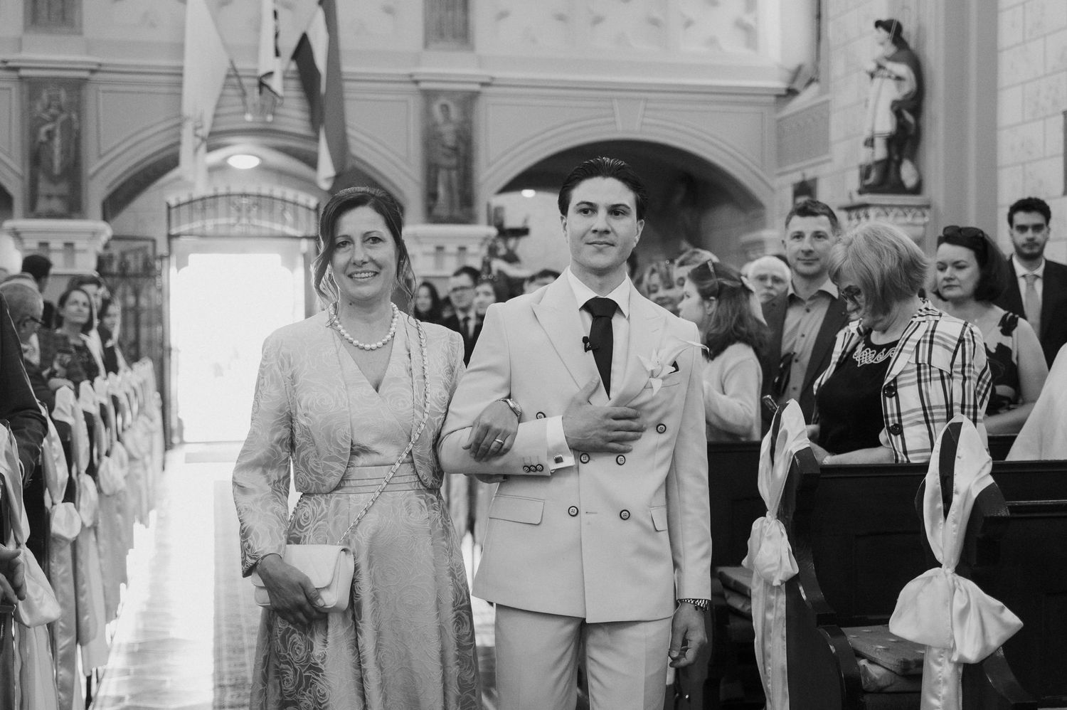 Groom escorted by mother down church aisle during traditional wedding ceremony in black and white photo.