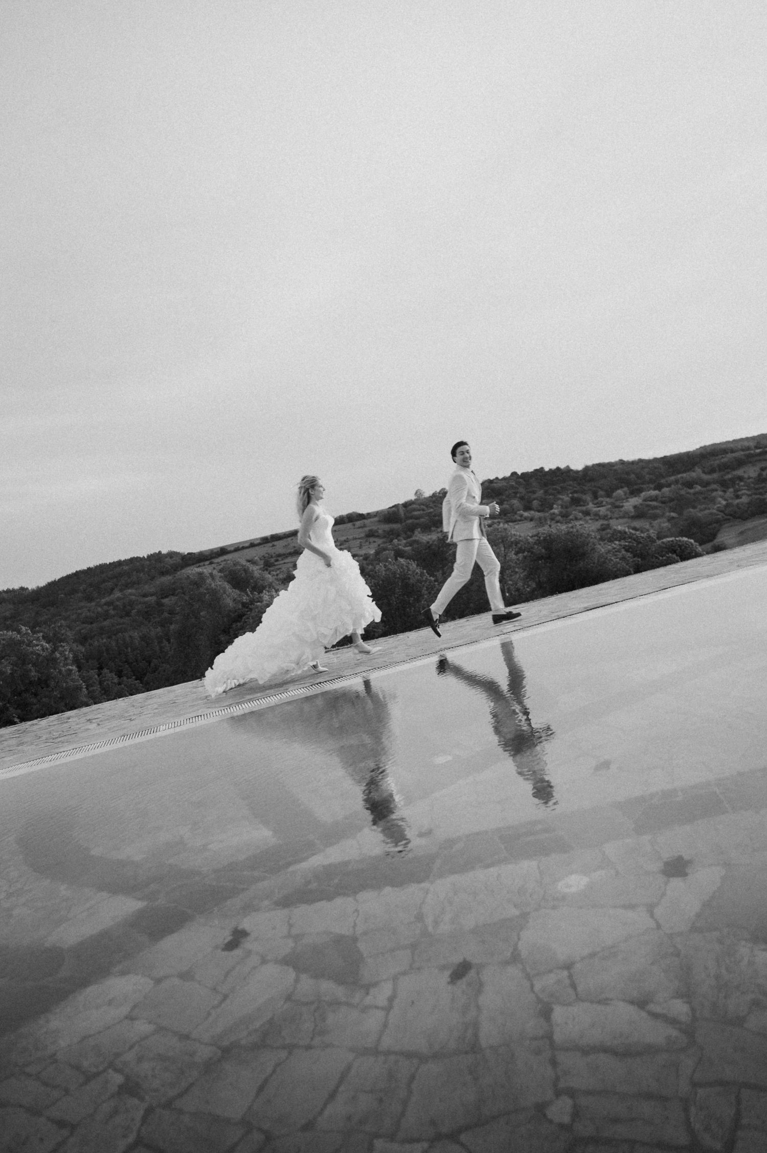 Black and white photo of bride and groom running beside a reflective pool with scenic hills in the background.