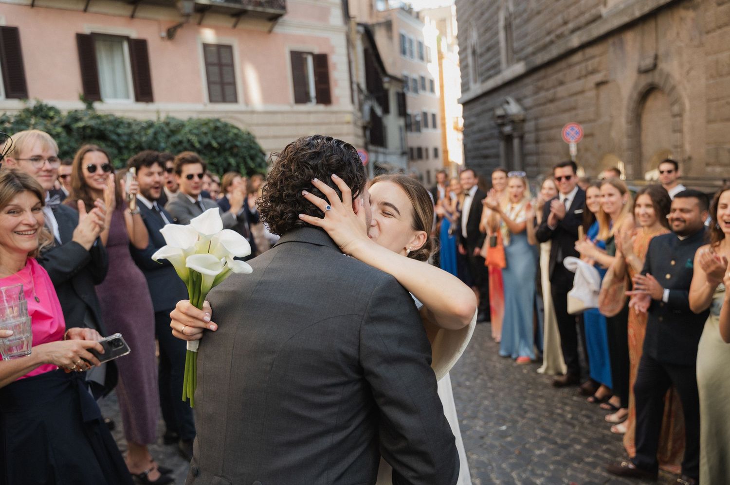 A romantic wedding kiss on a cobblestone street in Italy as guests cheer and celebrate.