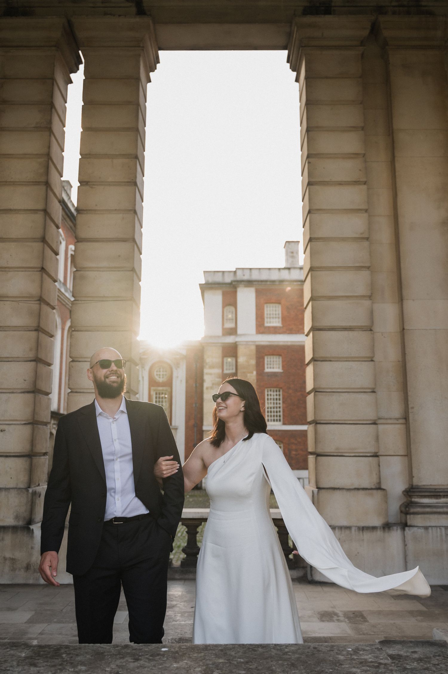 A couple in formal attire poses beneath classical architecture during sunset.