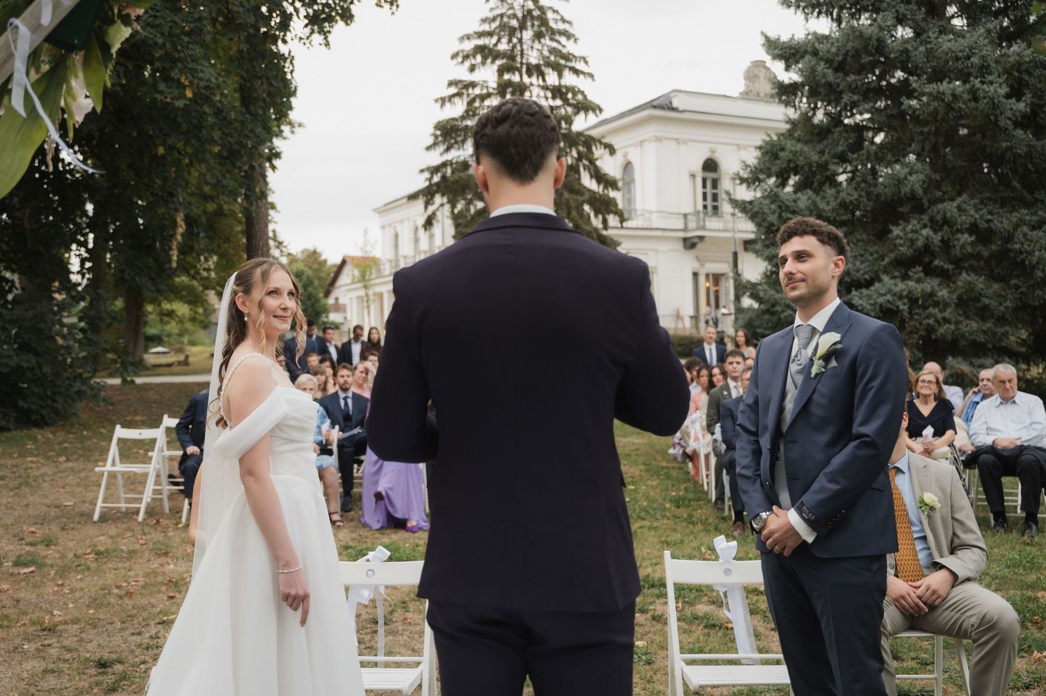 Outdoor wedding ceremony with bride in white gown exchanging vows before officiant and guests.