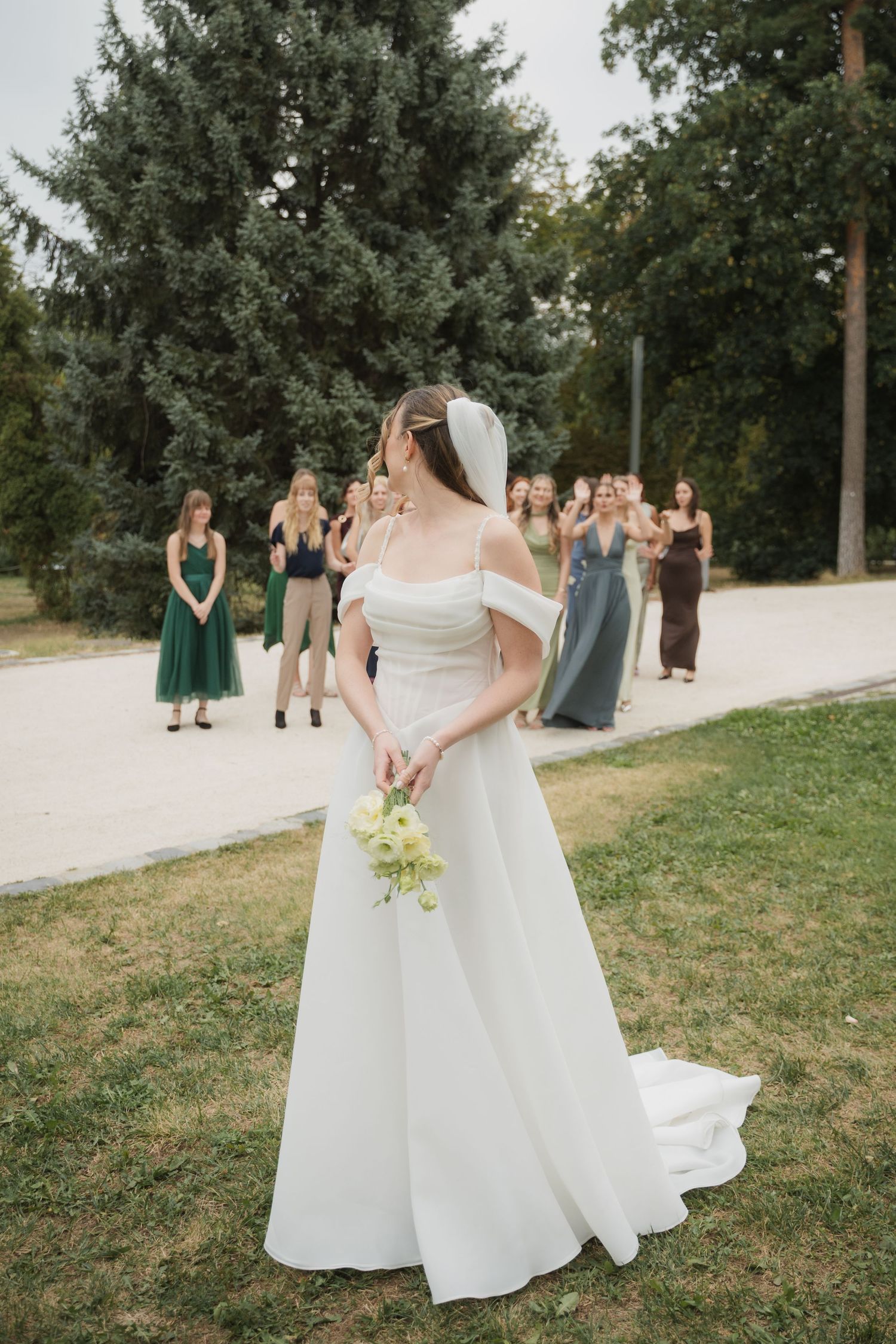 Bride in off-shoulder gown prepares to toss bouquet to excited guests gathered on garden lawn.