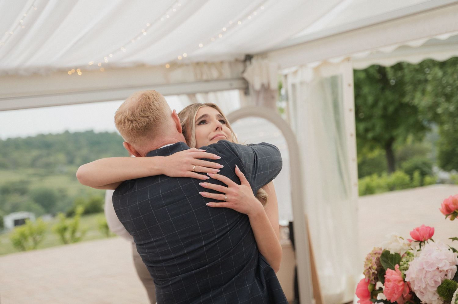 Bride and guest sharing warm embrace under marquee tent with floral arrangements at wedding reception.