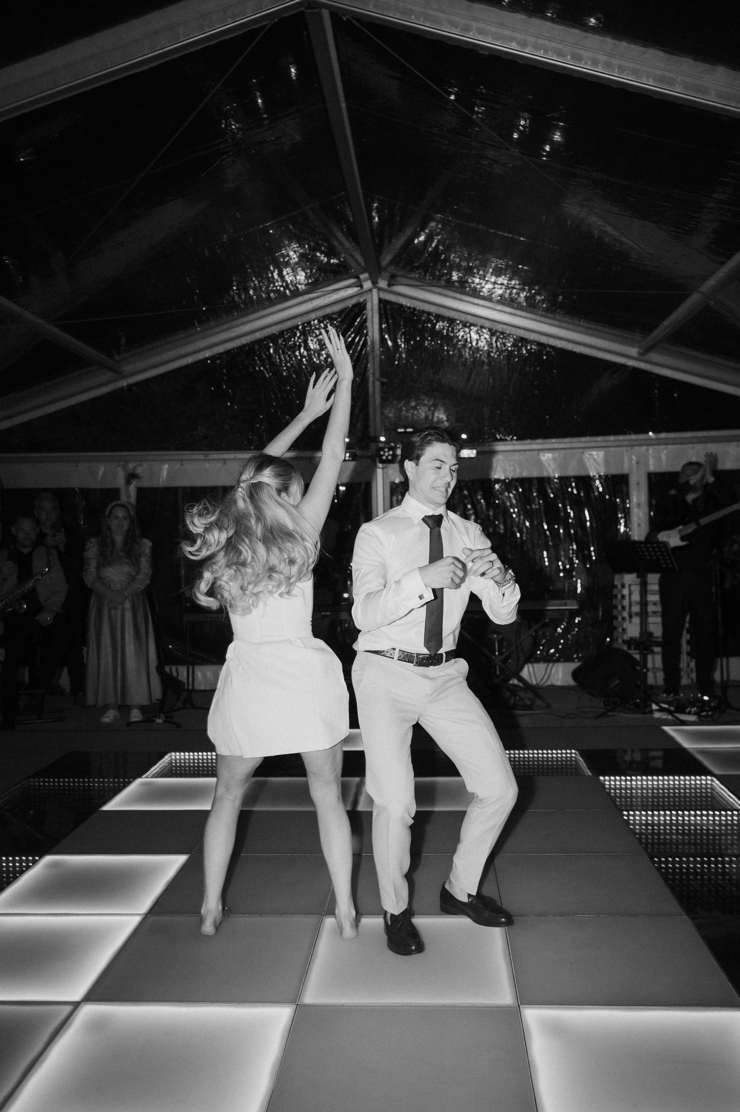 Black and white photo of bride and groom dancing energetically on a lit dance floor at wedding reception.