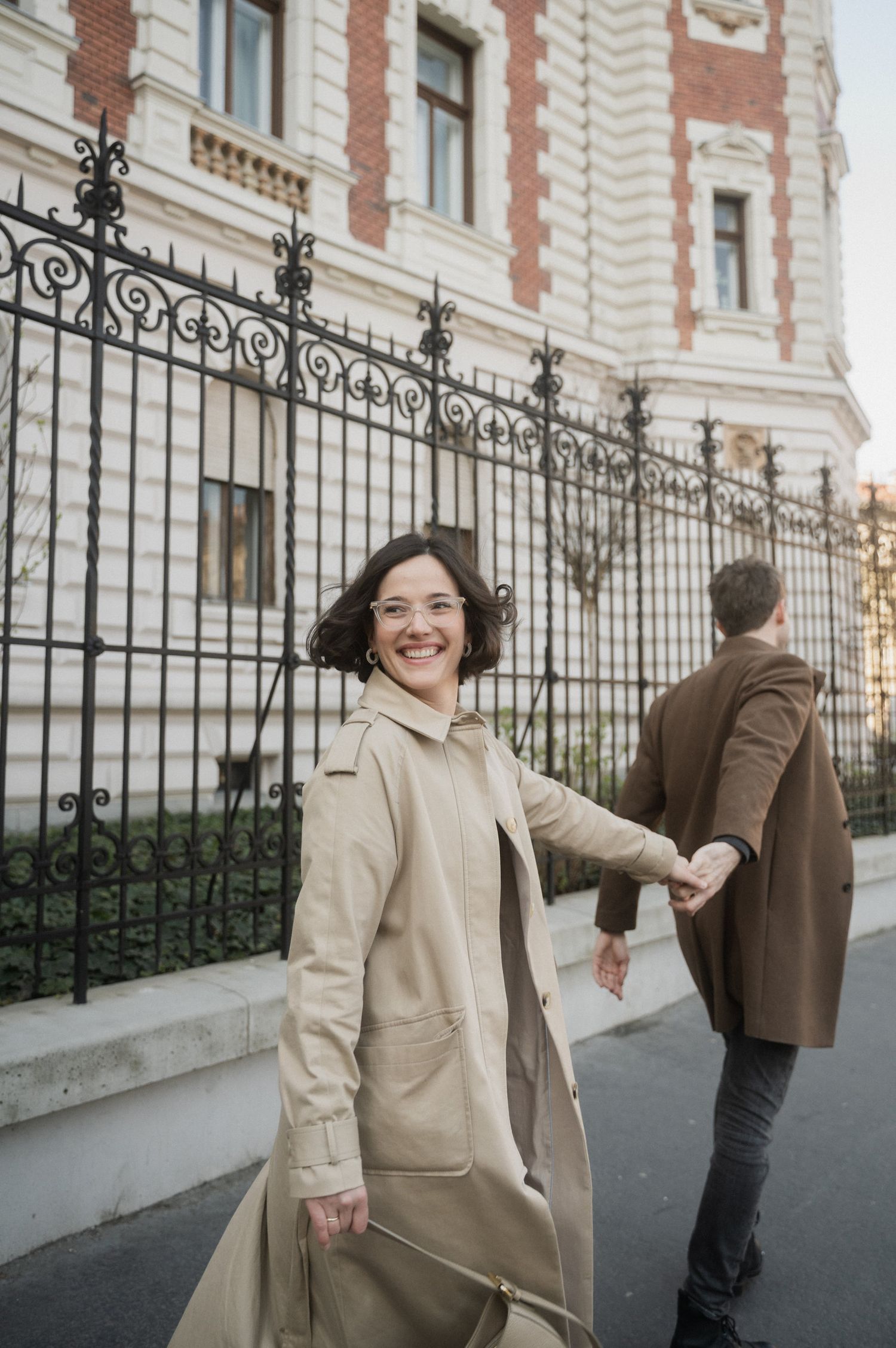 A couple in vintage style clothing shares playful moments together along an ornate iron fence outside a brick building.