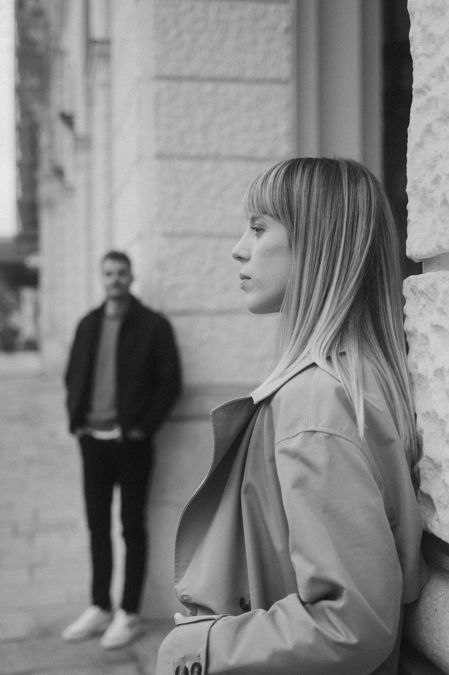Black and white portrait of two people in an urban setting with one person in a trench coat against a stone wall.