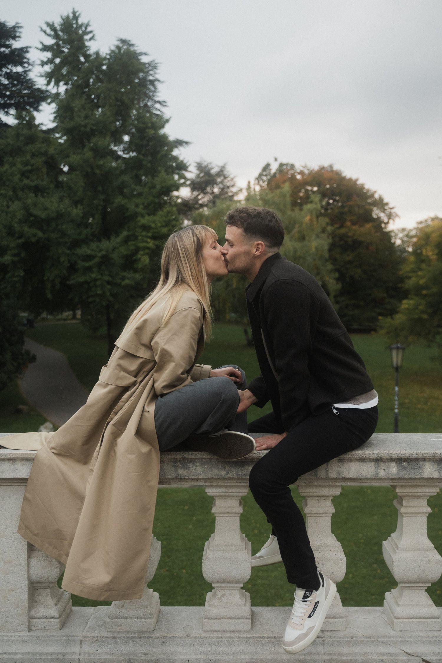 A romantic couple sits on a stone balustrade in a park, sharing an intimate moment while wearing casual attire.