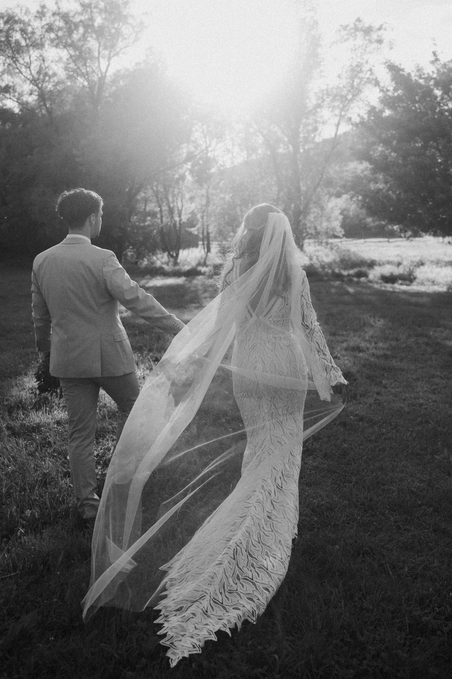 A romantic black and white wedding photo series showing a couple walking through a sunlit field with a flowing veil in the breeze.