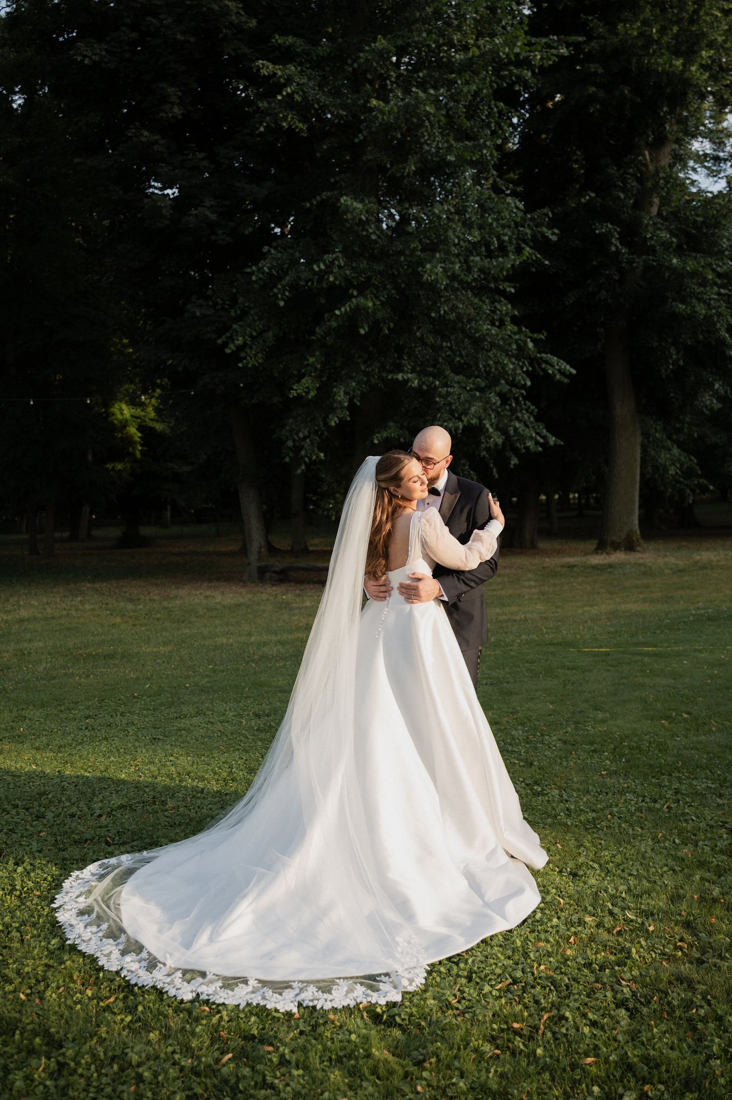 A romantic wedding couple embraces on a grassy lawn with flowing white dress and veil trailing behind them.