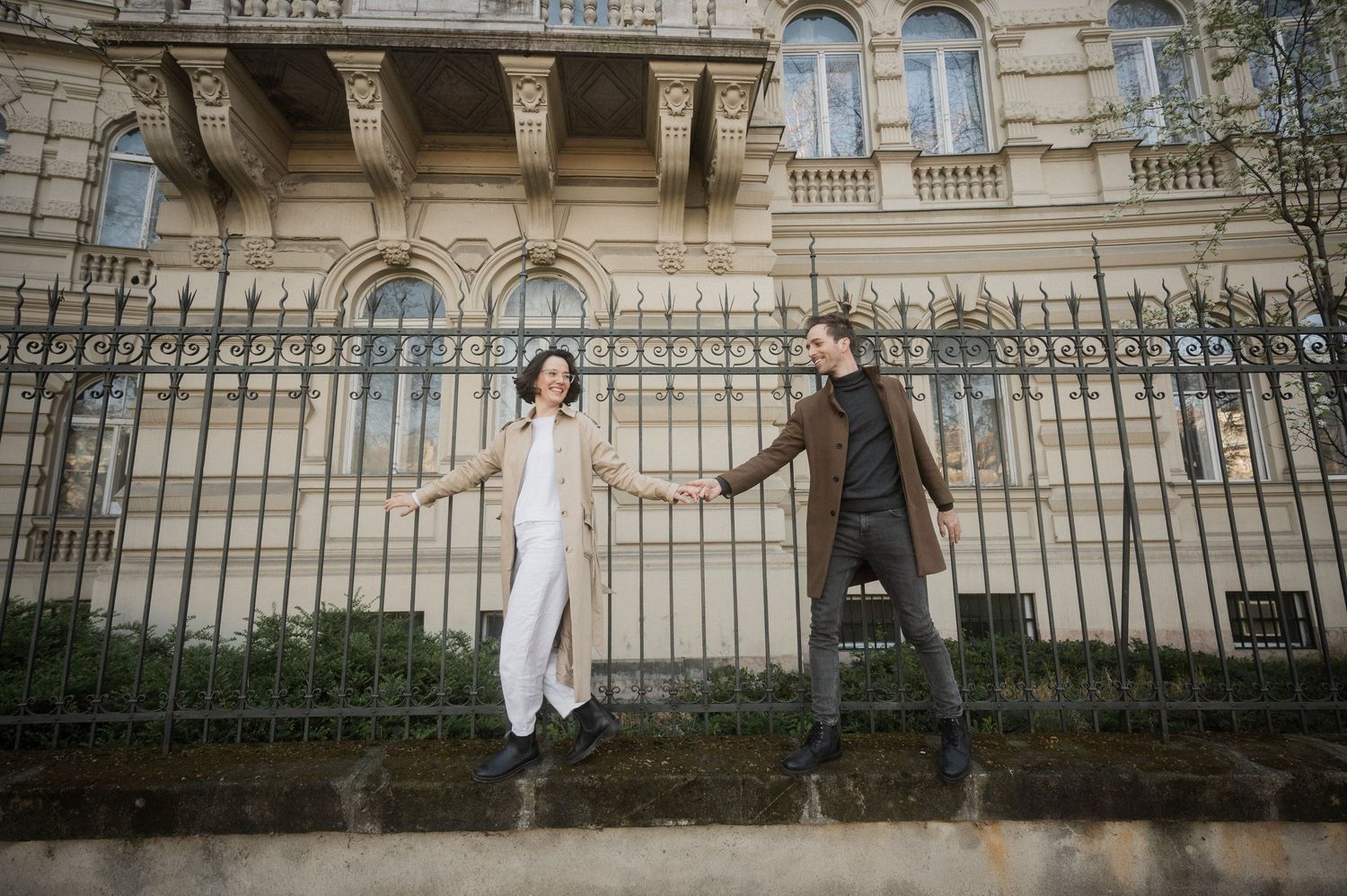 Two people in contrasting outfits jumping in sync in front of an ornate historic building with iron gates.