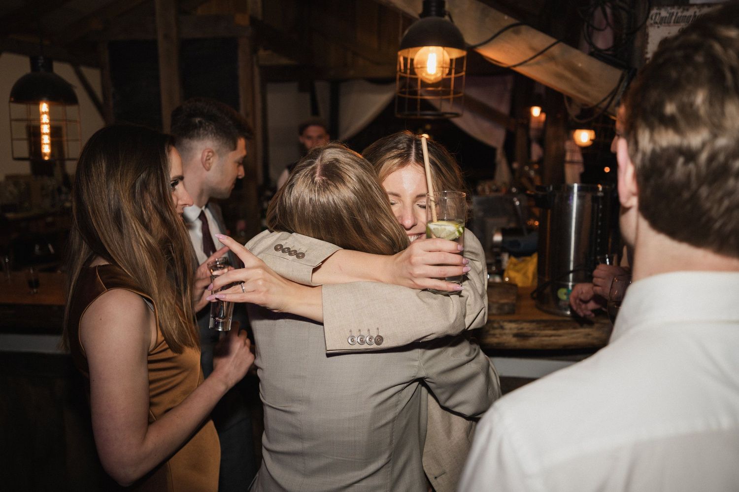 People dancing and celebrating at a dimly lit party venue with hanging pendant lights.