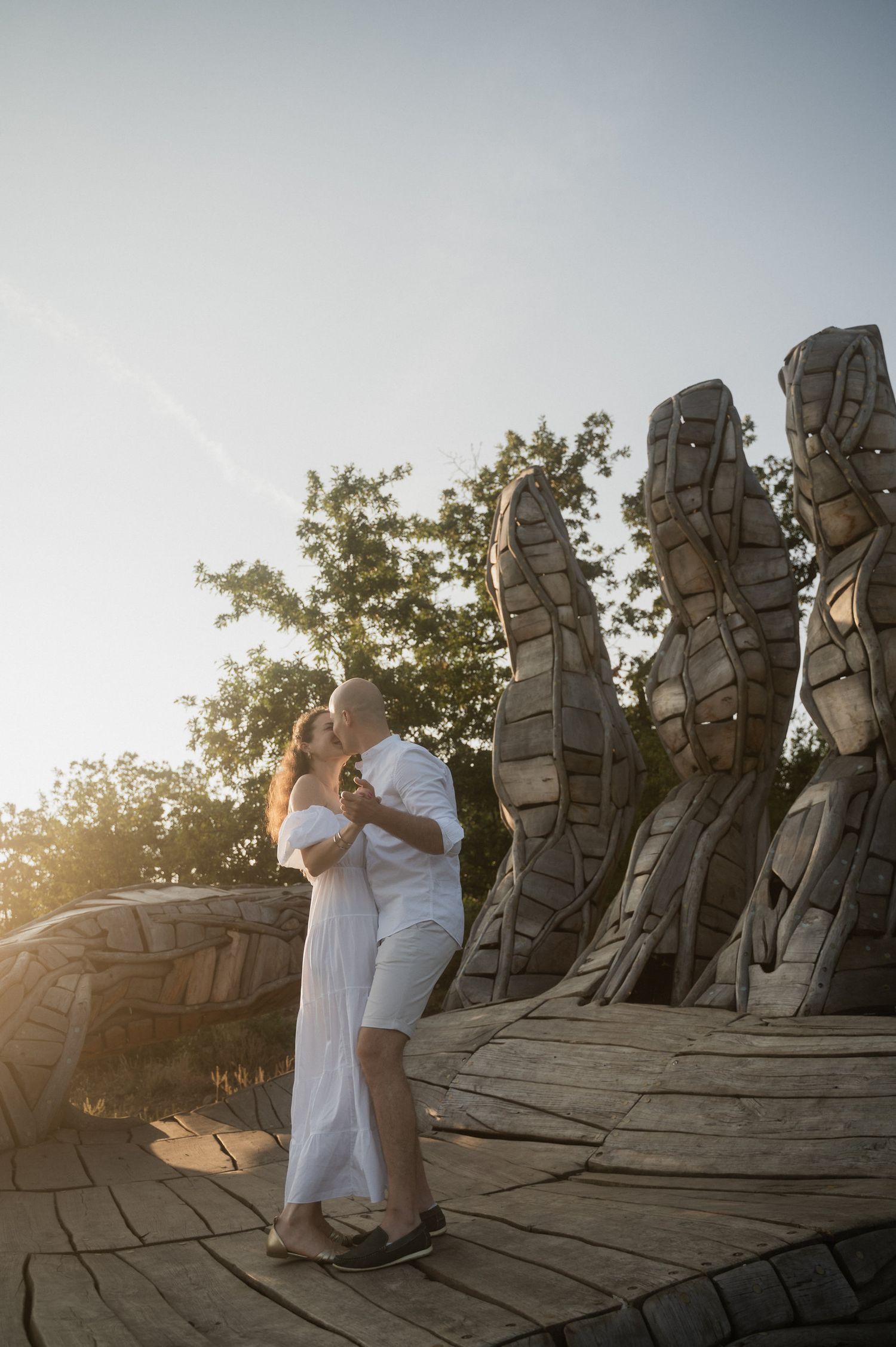 A couple shares an intimate moment at sunset against a backdrop of stone sculptures in an outdoor setting.