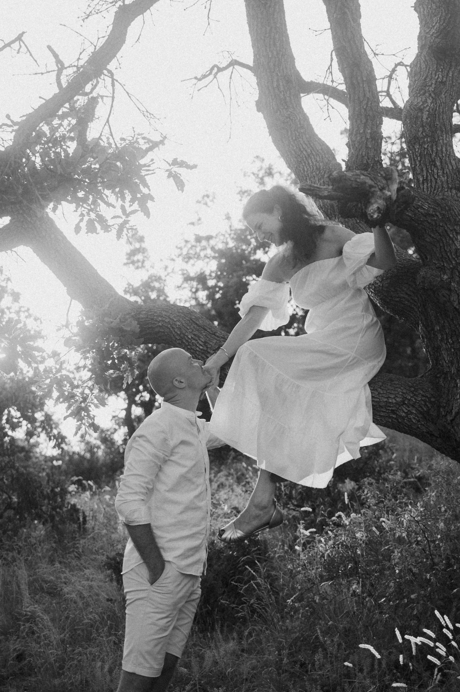 Two people in white clothing share a romantic dance pose against a sunlit wooded backdrop.