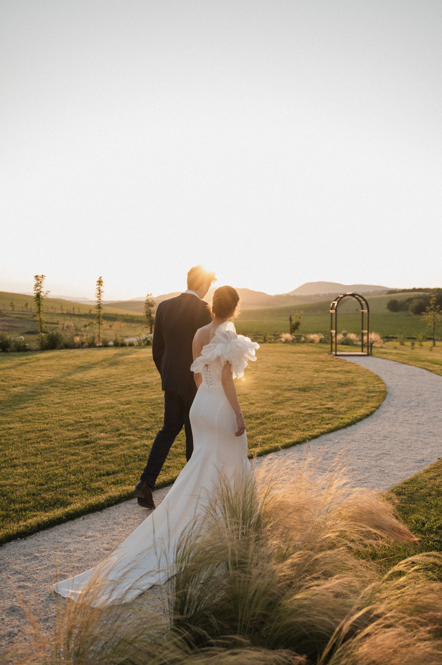 A couple in wedding attire walks along a winding path at sunset with golden grasses swaying in the foreground.