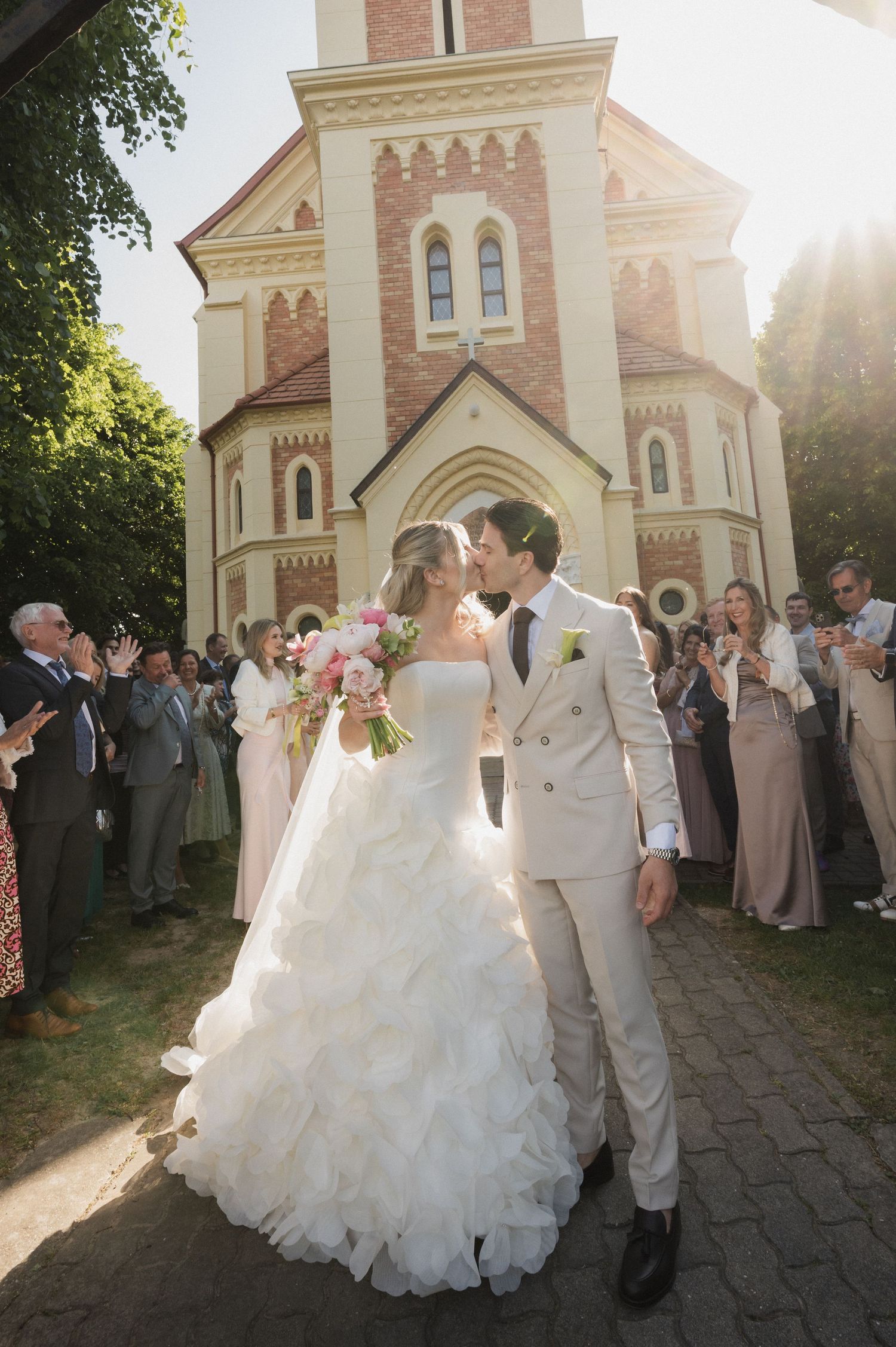 Newlyweds share a kiss outside a historic church surrounded by applauding wedding guests on a sunny day.