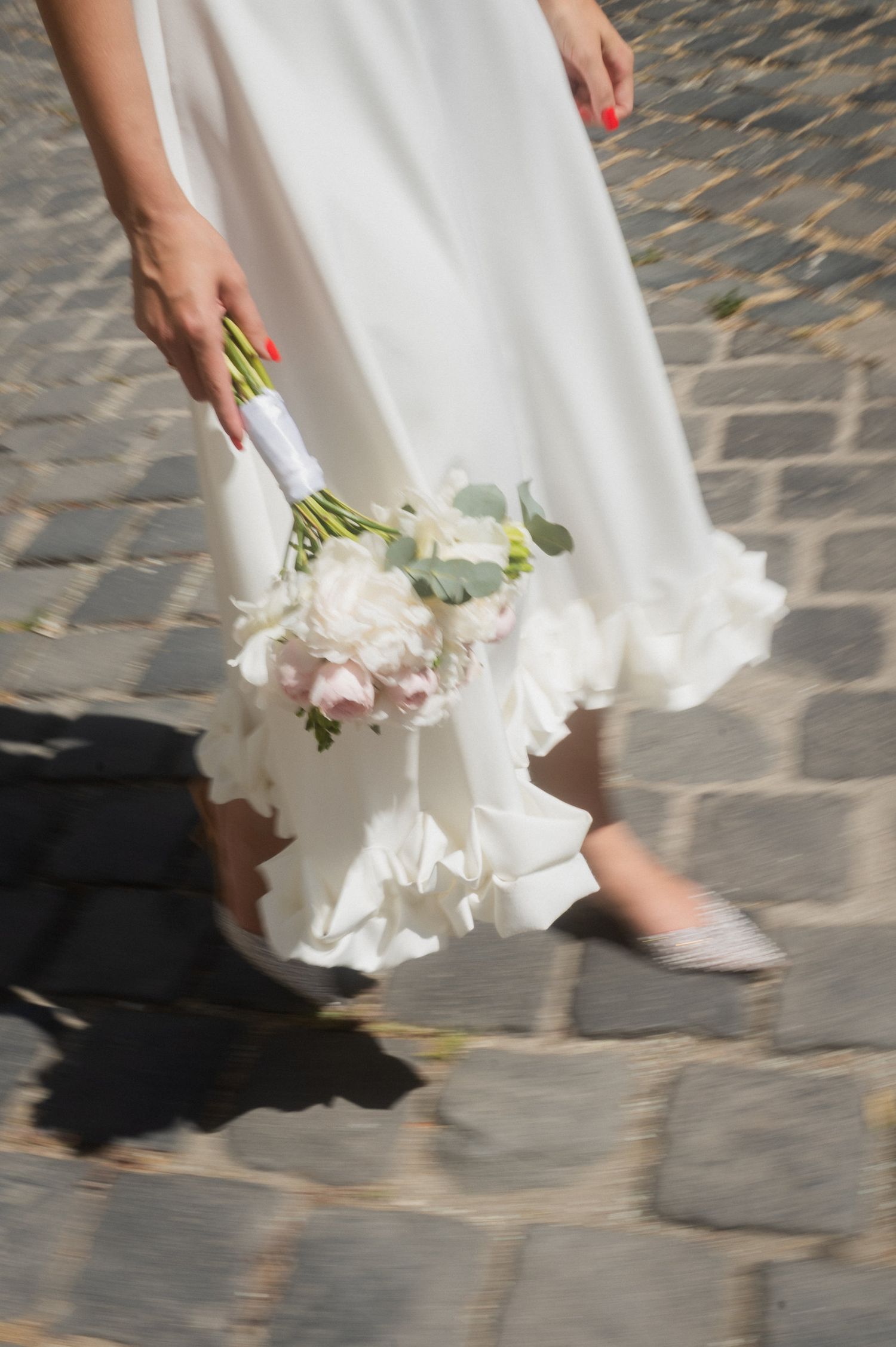 Close-up of white wedding dress hem and bouquet against cobblestone ground.