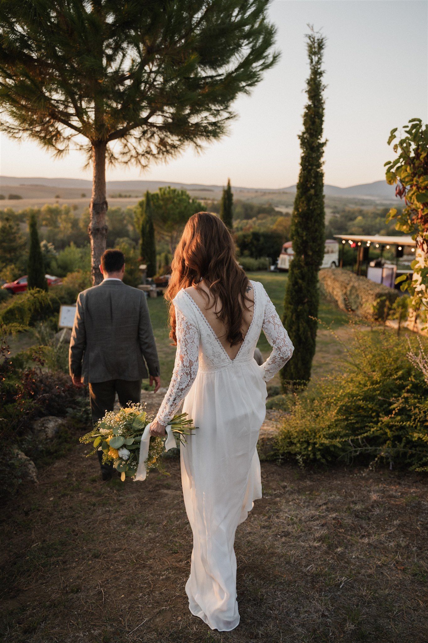Bride in long-sleeved lace gown holding sunflowers walks through Tuscan-style garden at sunset.
