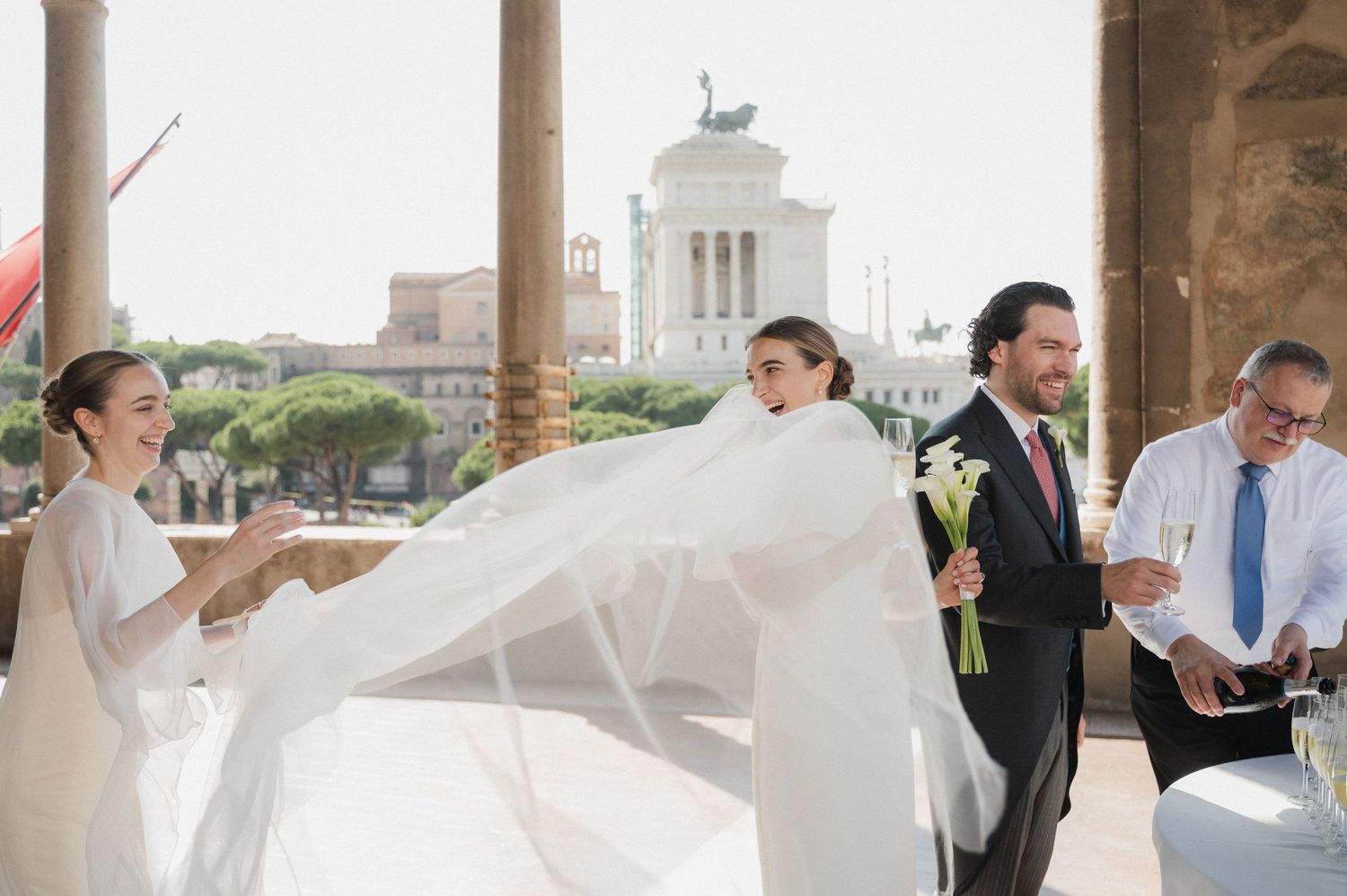 Wedding party gathers under stone archways with a historic cityscape backdrop.