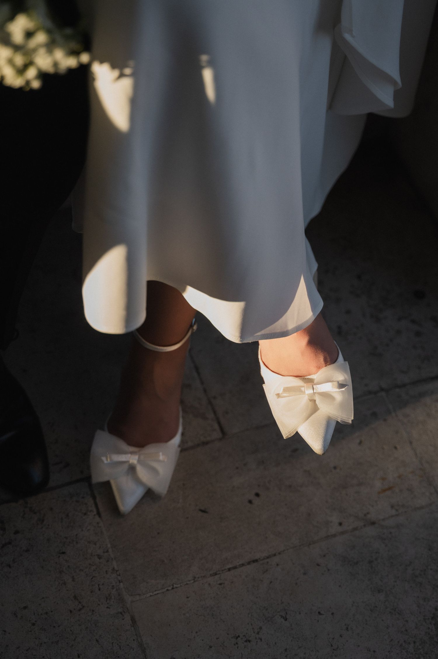 Detail shot of elegant white bridal shoes and dress hem on stone flooring.