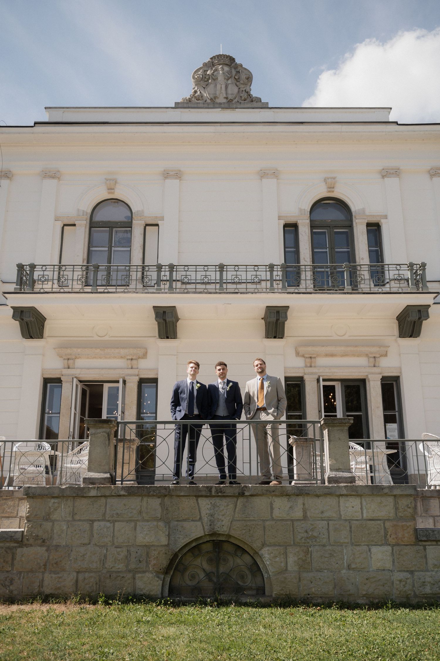 Three groomsmen in suits stand on an elegant white villa balcony with iron railings under a partly cloudy sky.