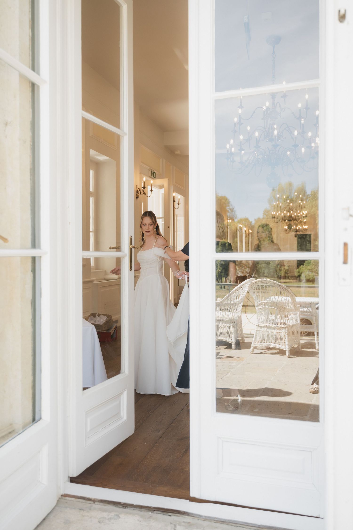 Bride in white wedding gown standing in elegant French doors, with wicker chairs visible in sunny outdoor reflection.