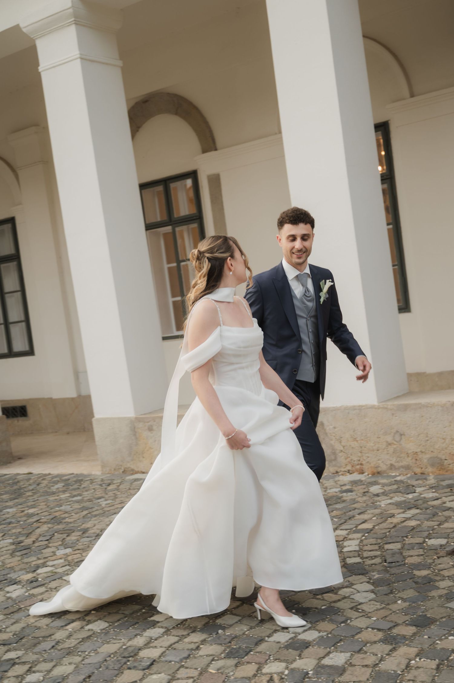 Bride in off-shoulder white gown and groom in navy suit walking hand in hand.