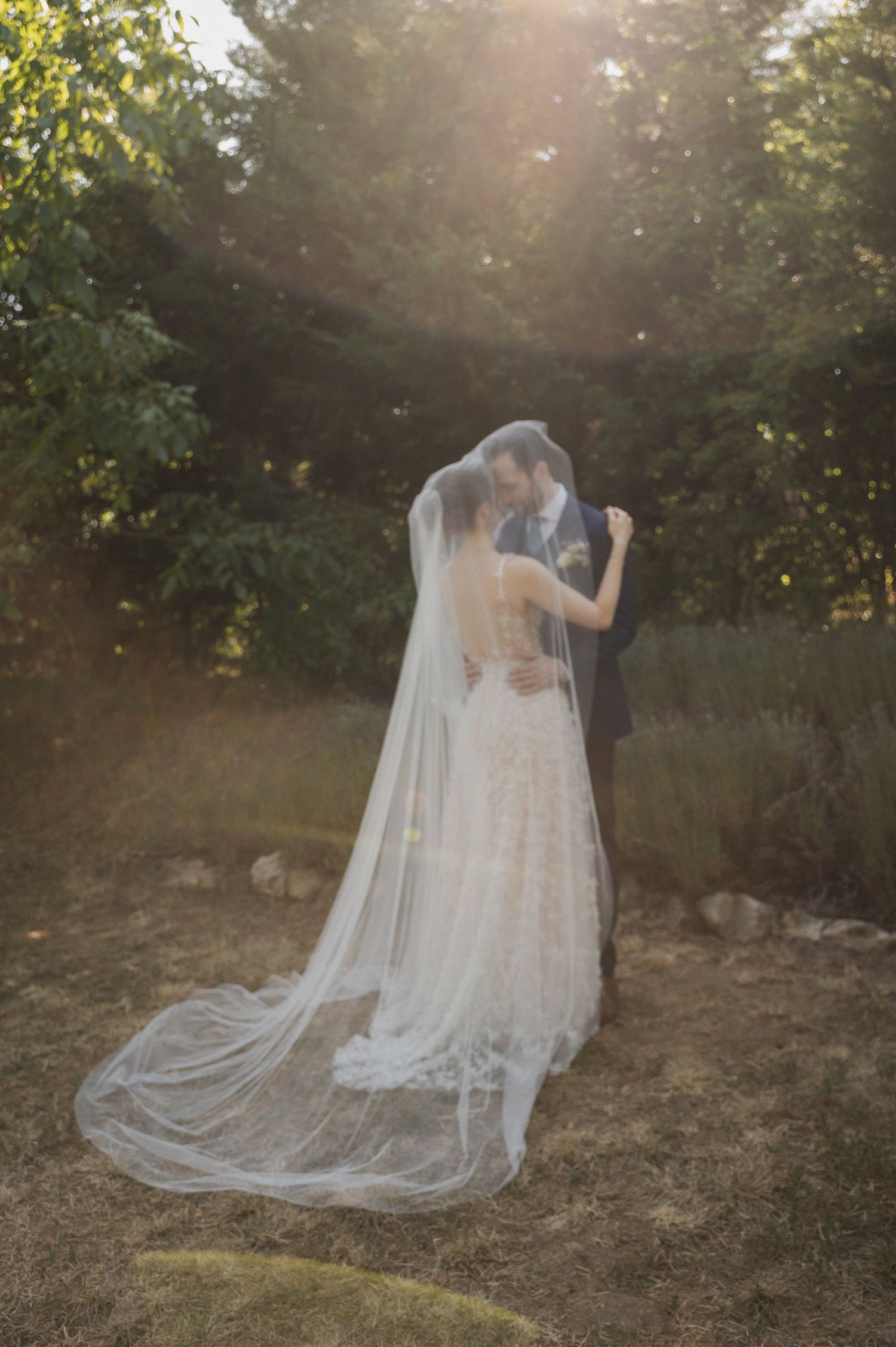 A romantic sunset wedding portrait shows a couple embracing with a flowing cathedral veil trailing behind them.