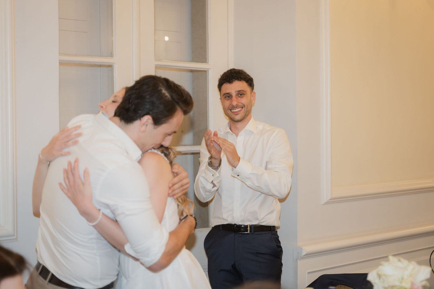 Bride and groom share a hug while smiling guest claps in elegant white reception hall during wedding celebration.