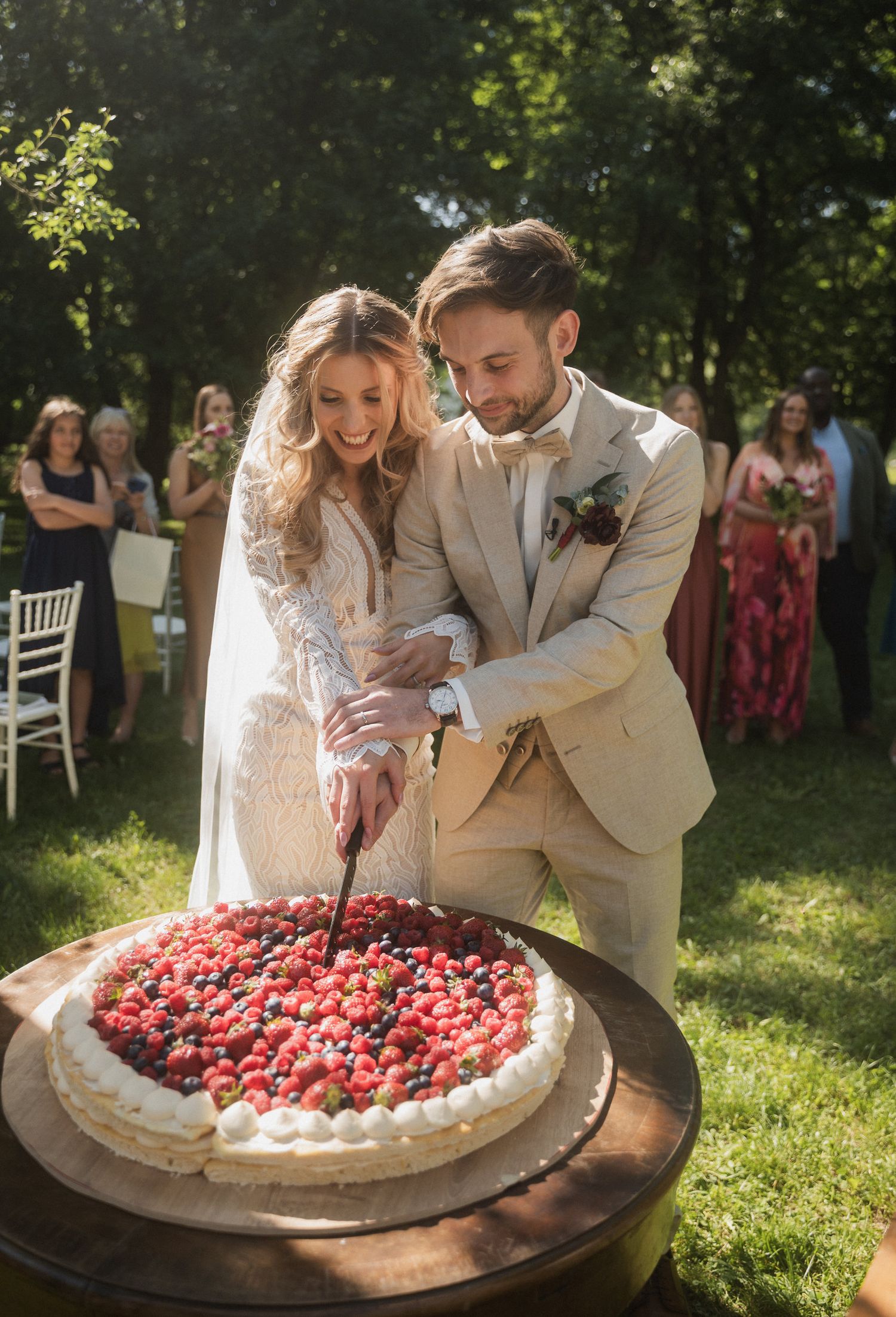 A couple in wedding attire cutting into a large pavlova dessert decorated with fresh strawberries at an outdoor reception.