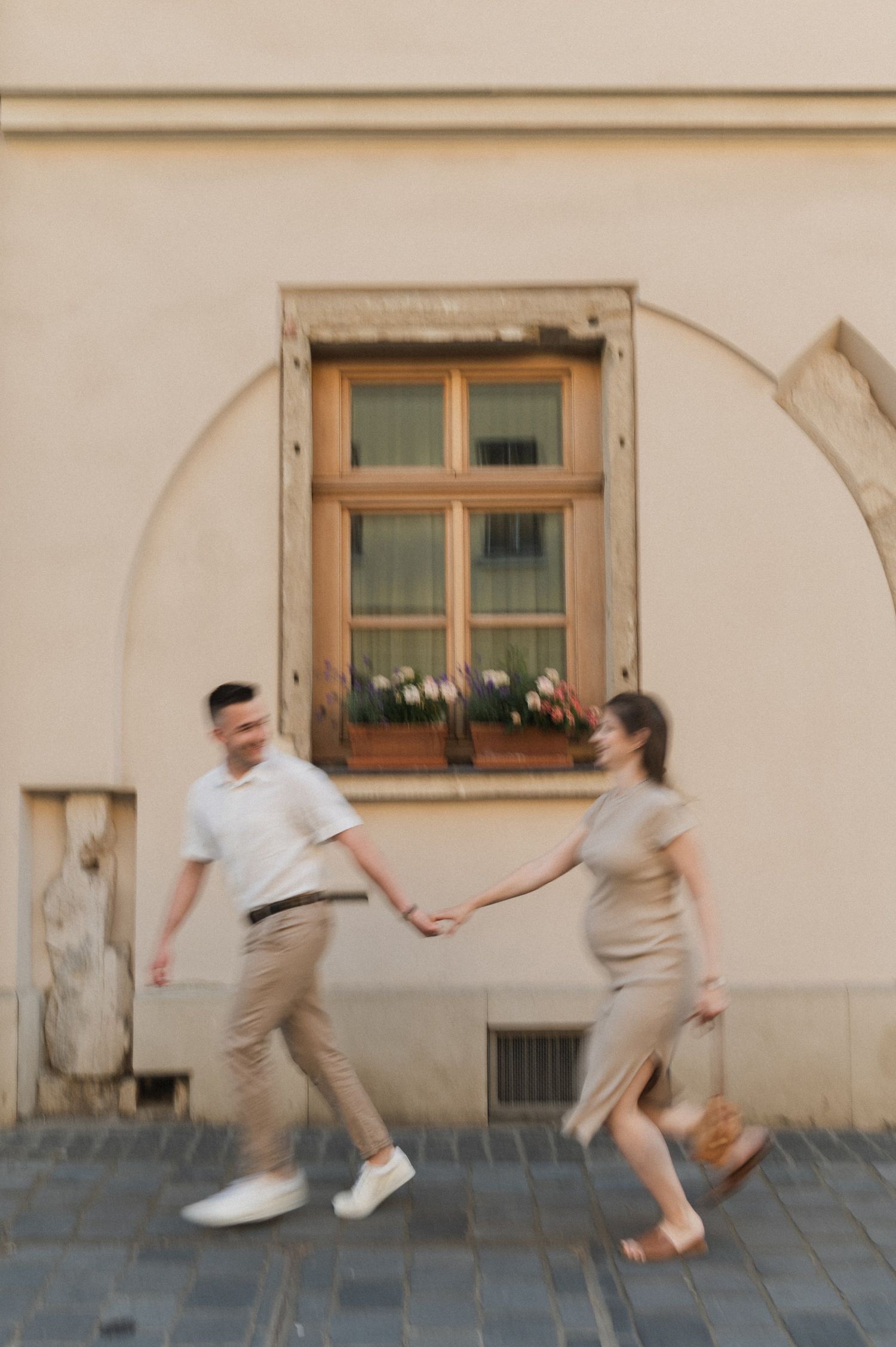 Two people playfully chase a basketball on a cobblestone street in front of a building with wooden windows.