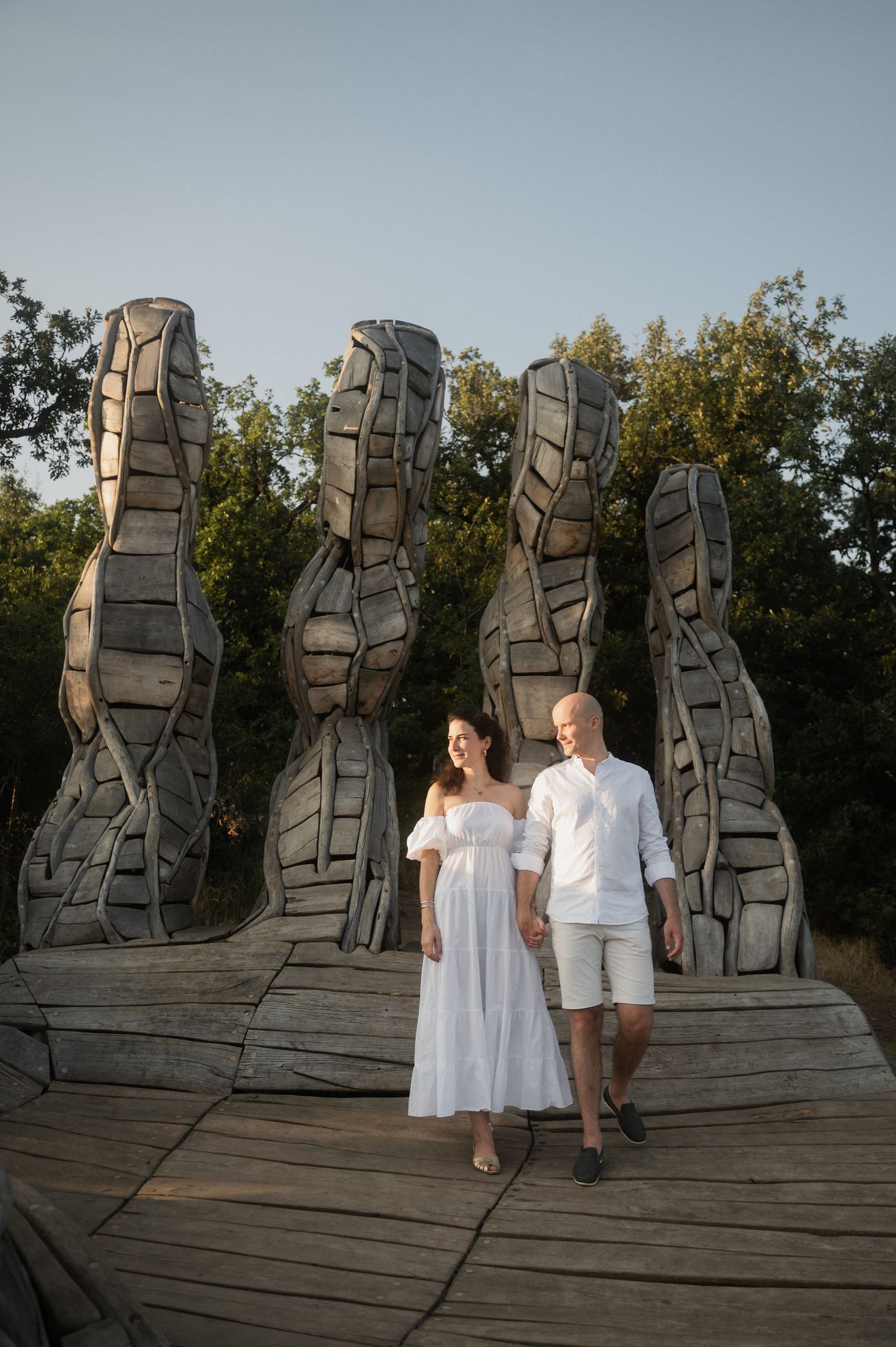 A couple in white outfits poses for a series of artistic photos in front of large abstract stone sculptures at sunset.