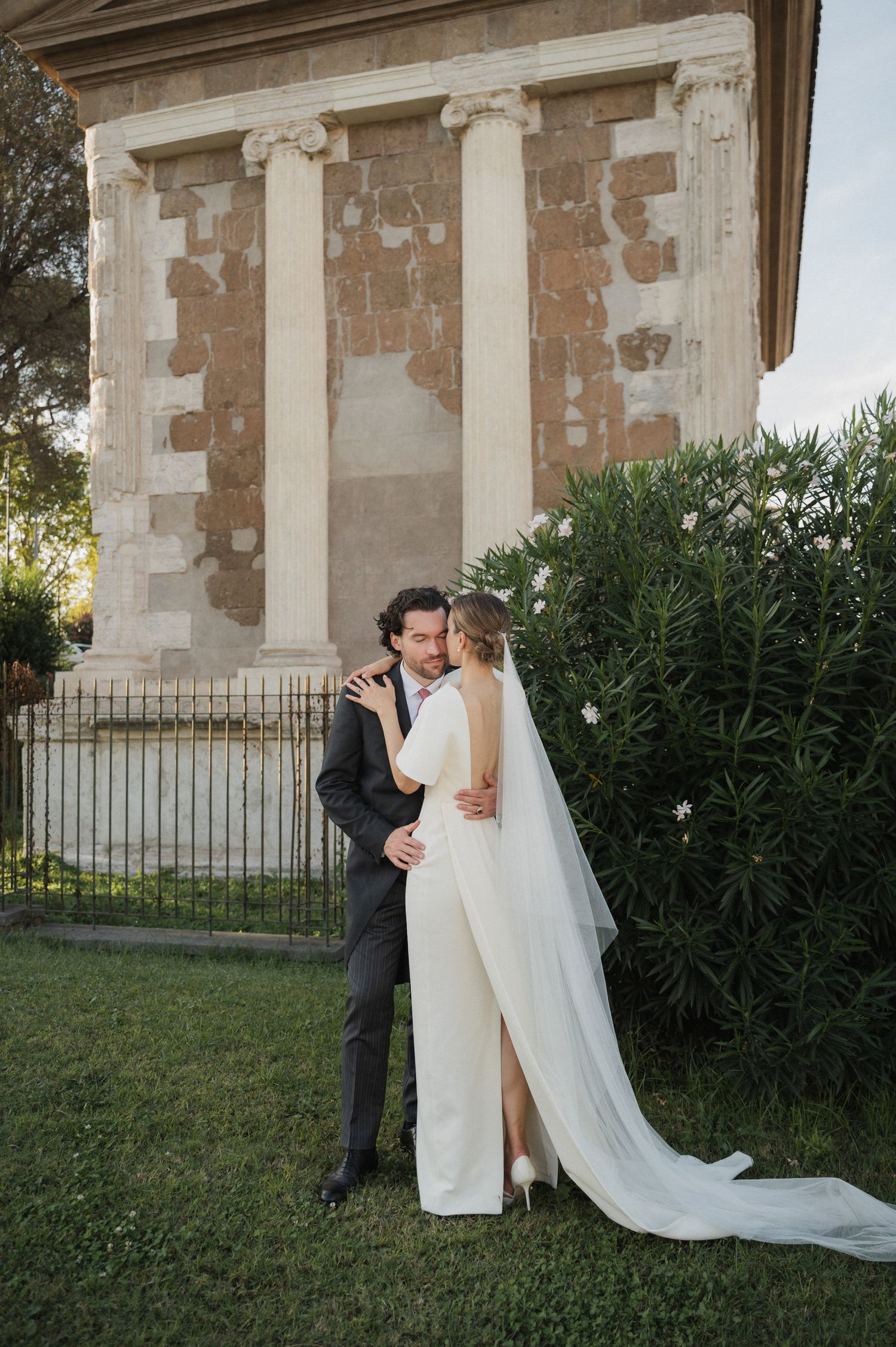 A wedding couple embraces near a classical stone building with columns.