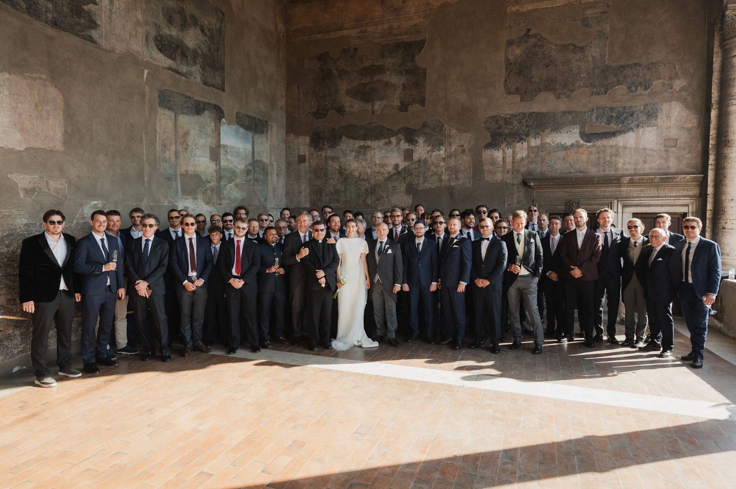 Large wedding party poses for a panoramic group photo in a historic stone venue.