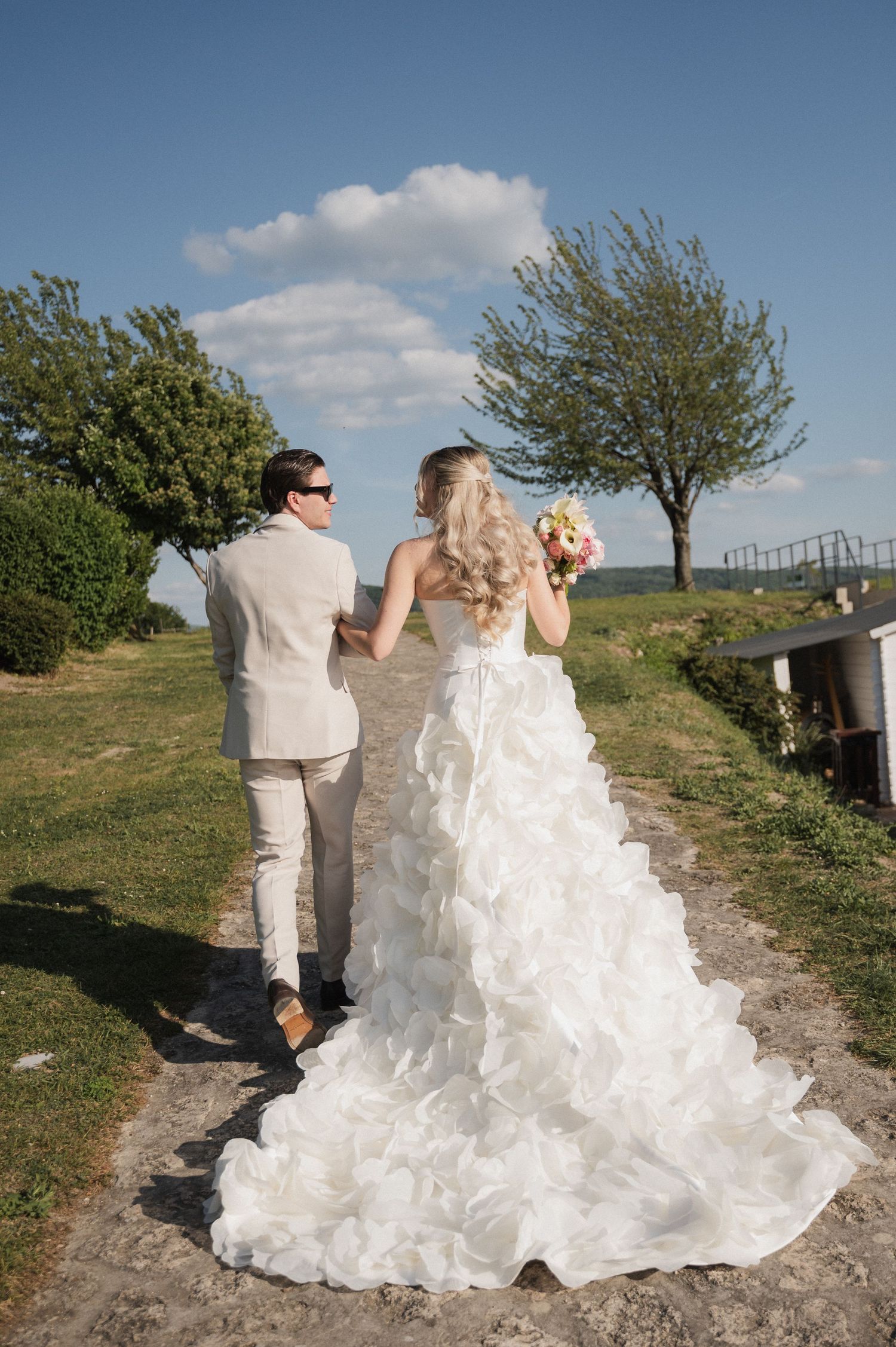 Bride and groom walk hand-in-hand down a scenic outdoor path, bride wearing a ruffled white gown holding a bouquet.