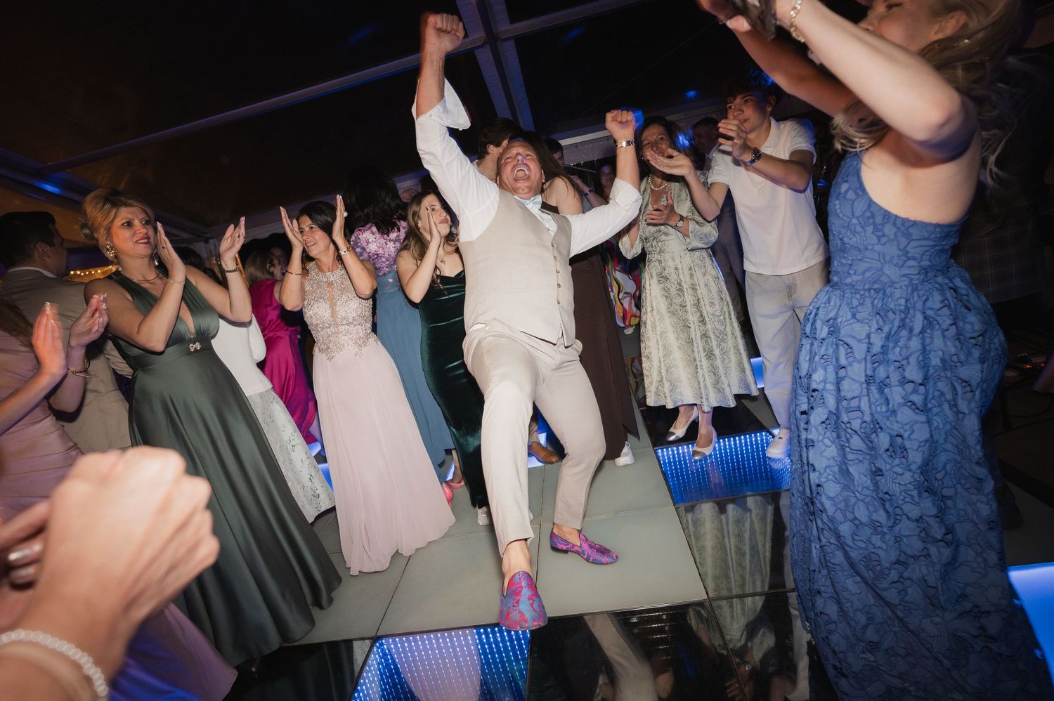 Excited wedding guests and groom dancing and jumping together on a glowing LED dance floor.