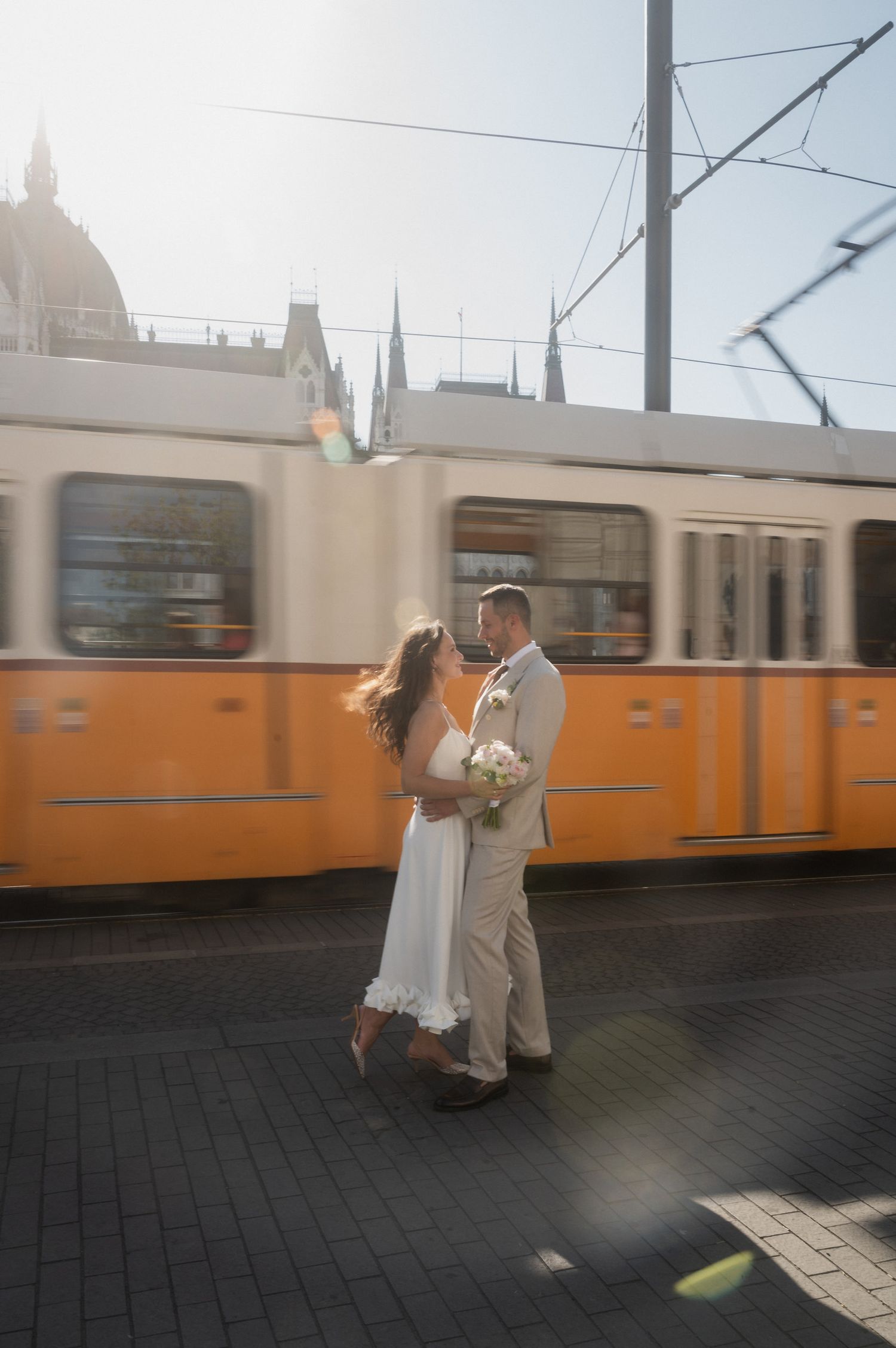 A couple embraces on a street with an orange tram passing in the background.