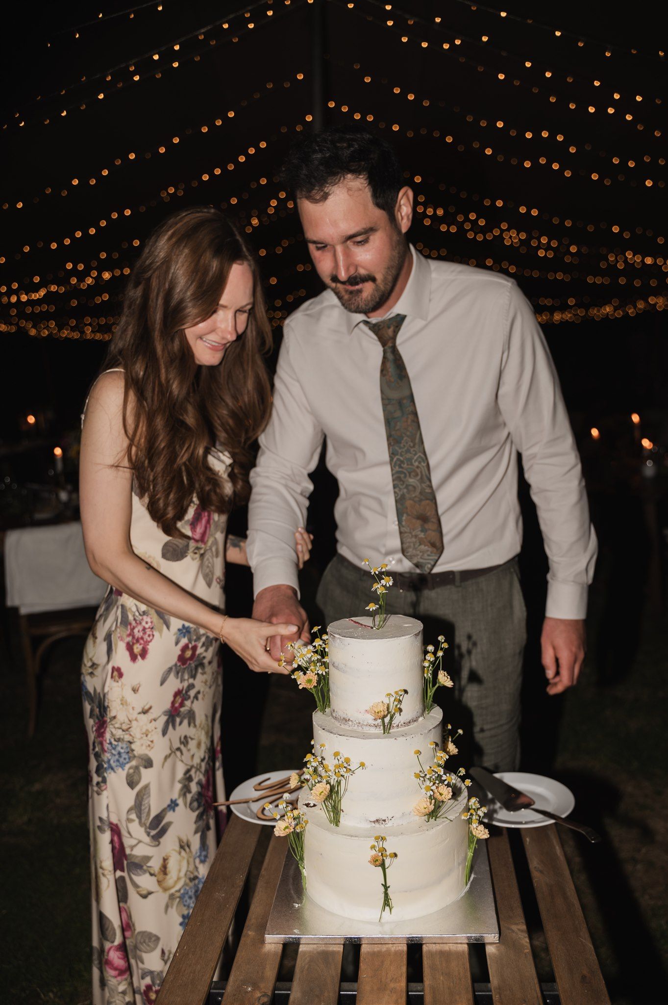 Couple cutting their elegant white wedding cake under string lights at night.