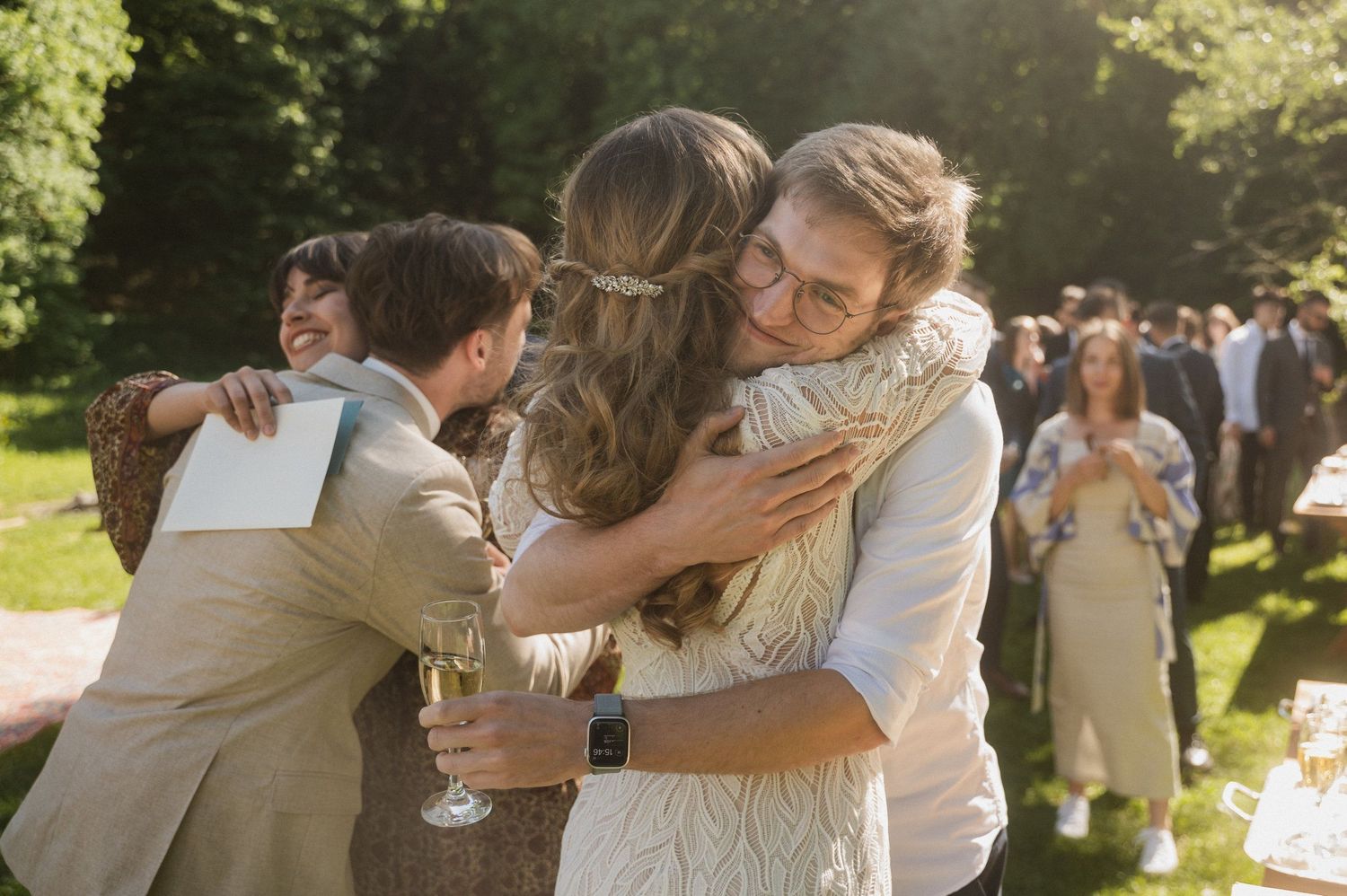 Wedding celebration in a sunlit garden with guests hugging and holding champagne glasses.