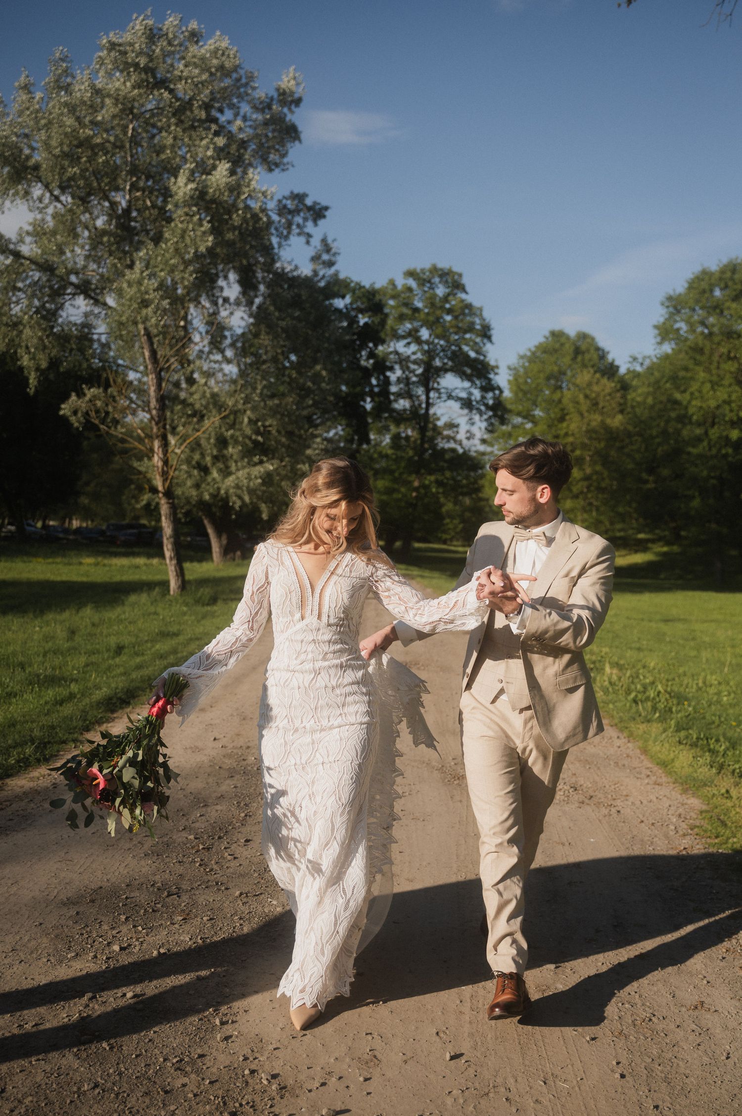 A couple in wedding attire dances playfully on a dirt path surrounded by green trees and grass on a sunny day.
