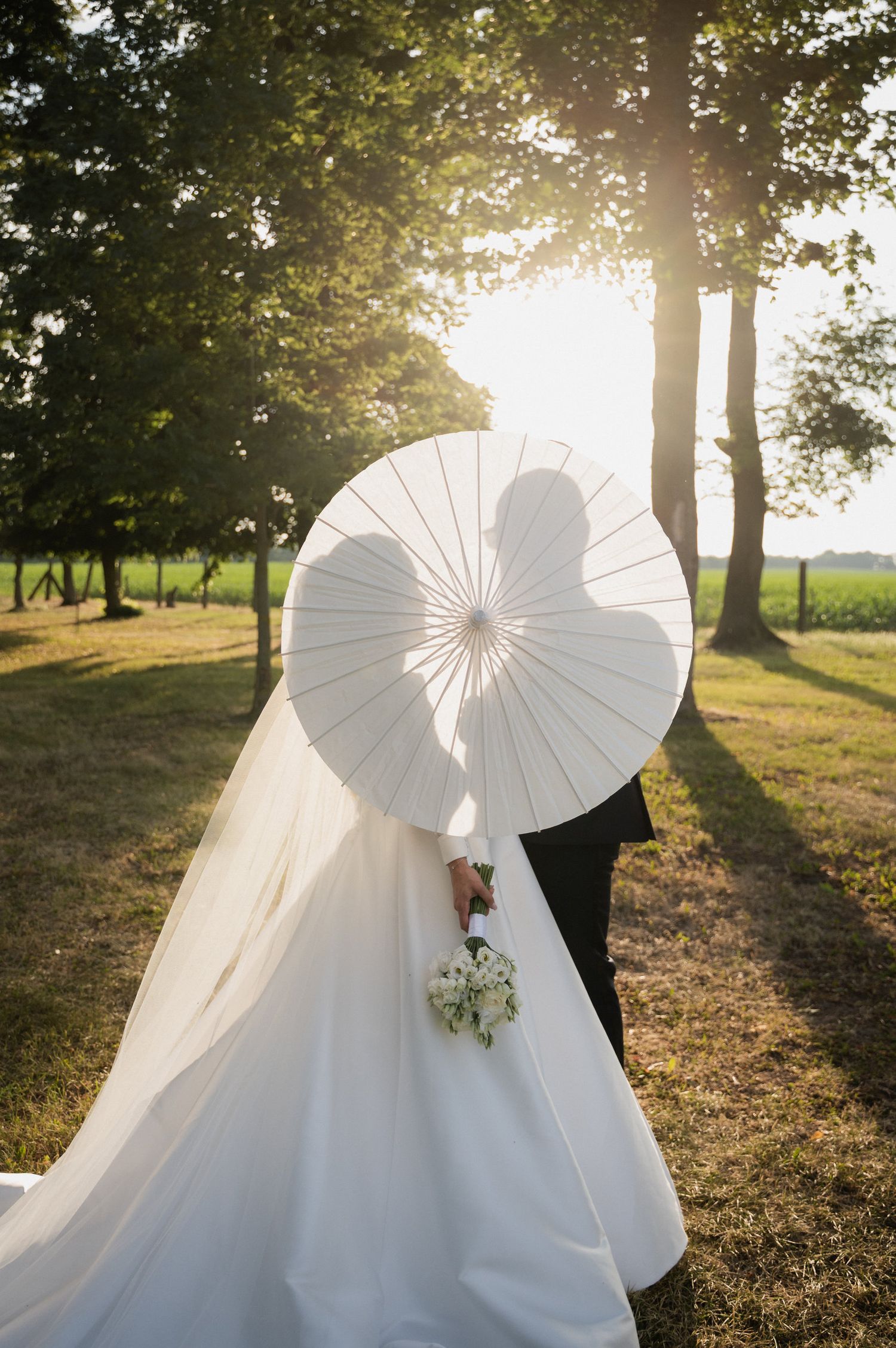 A romantic wedding scene with white parasols casting shadows in a sunlit park during golden hour.