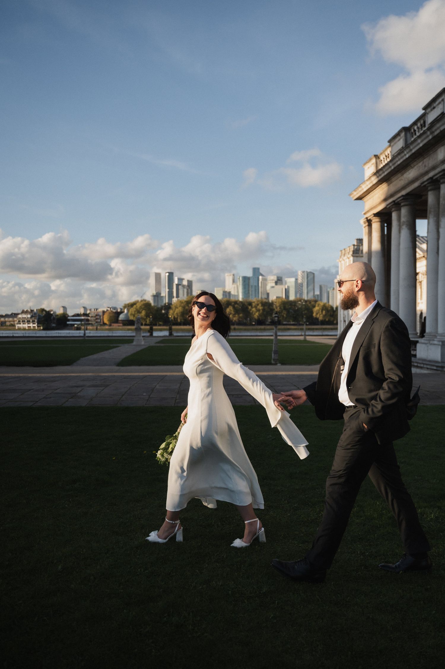 Playful wedding couple poses against dramatic city skyline at sunset.