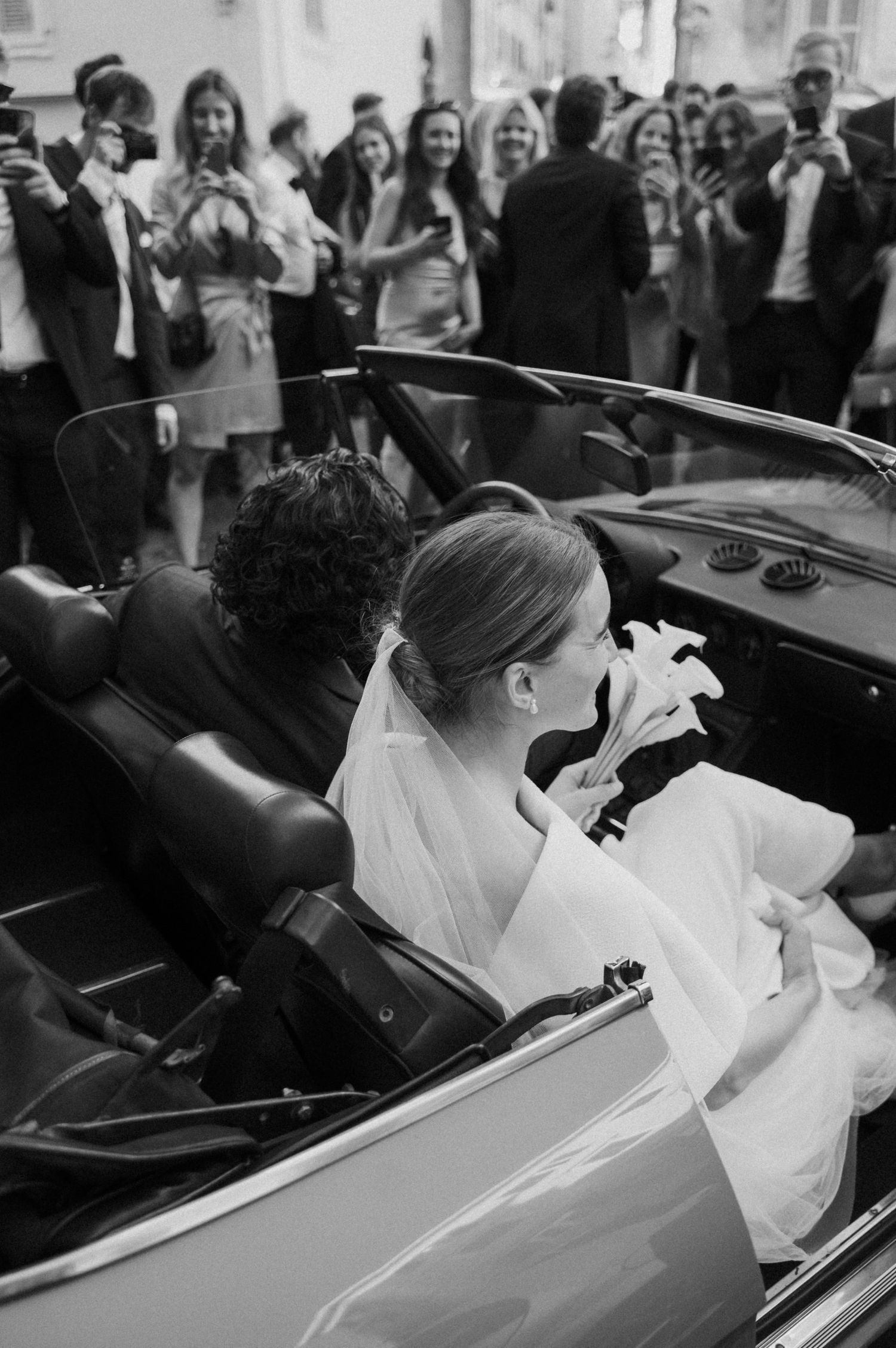 A black and white photo of newlyweds in a vintage convertible car surrounded by cheering guests.