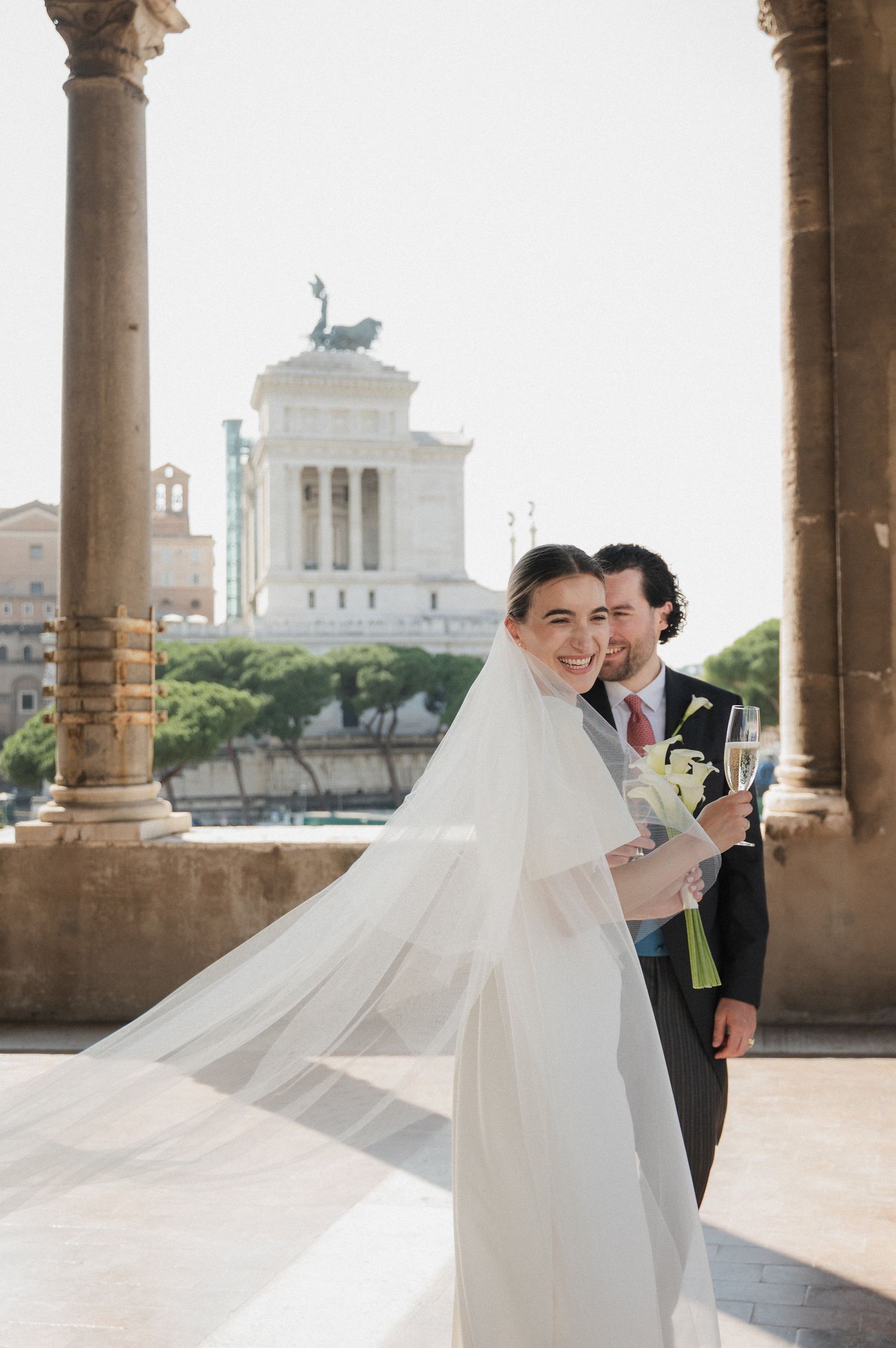Wedding couple under columned portico with Roman architecture and city views.