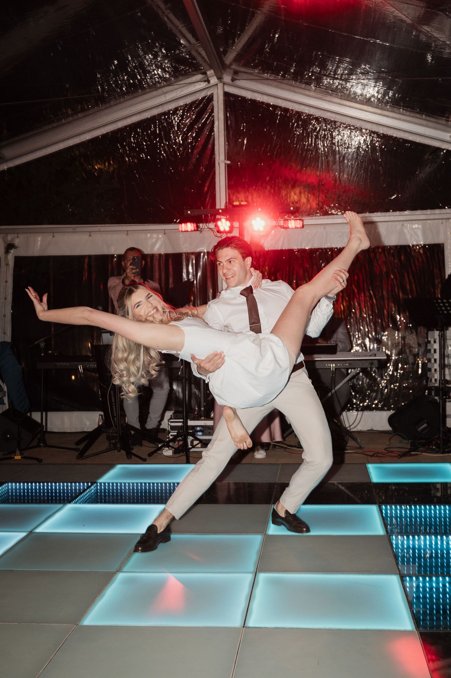 Groom dramatically lifts bride during energetic first dance performance on illuminated wedding dance floor.