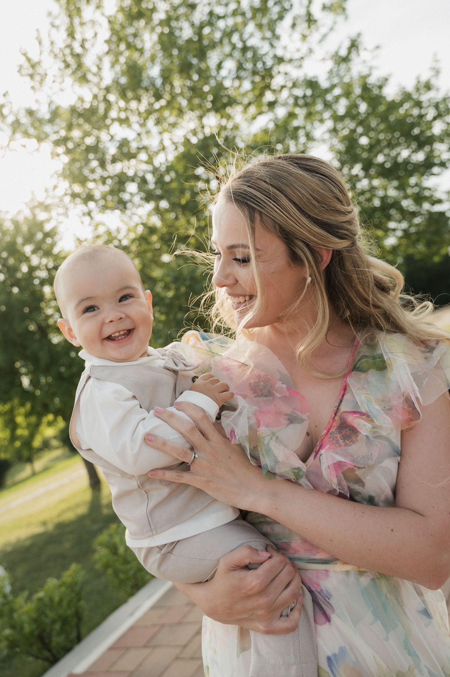A smiling mother in a floral dress holds her happy baby boy outdoors in a sunlit garden.