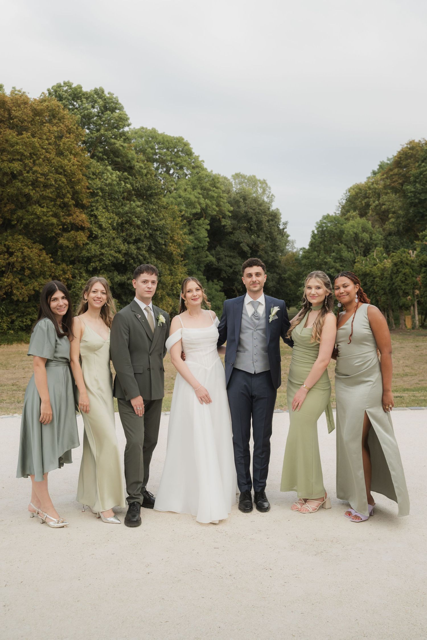 Wedding party group portrait with bride, groom, and bridesmaids in sage green dresses outdoors.