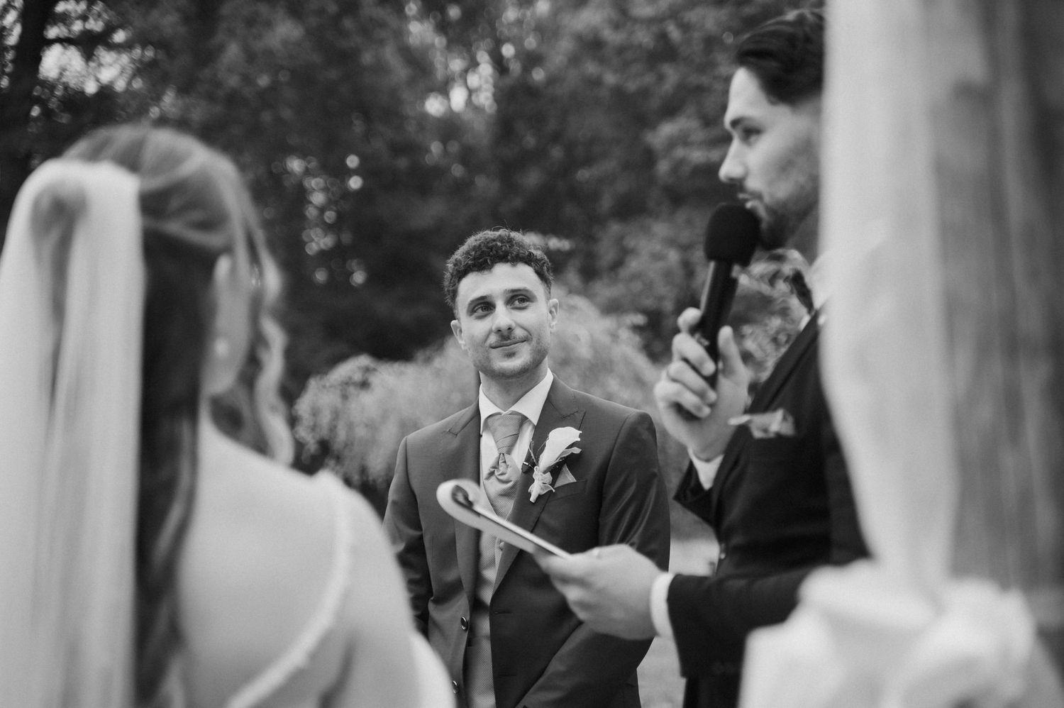 Groom smiling emotionally during outdoor wedding ceremony in black and white photo with officiant and bride.