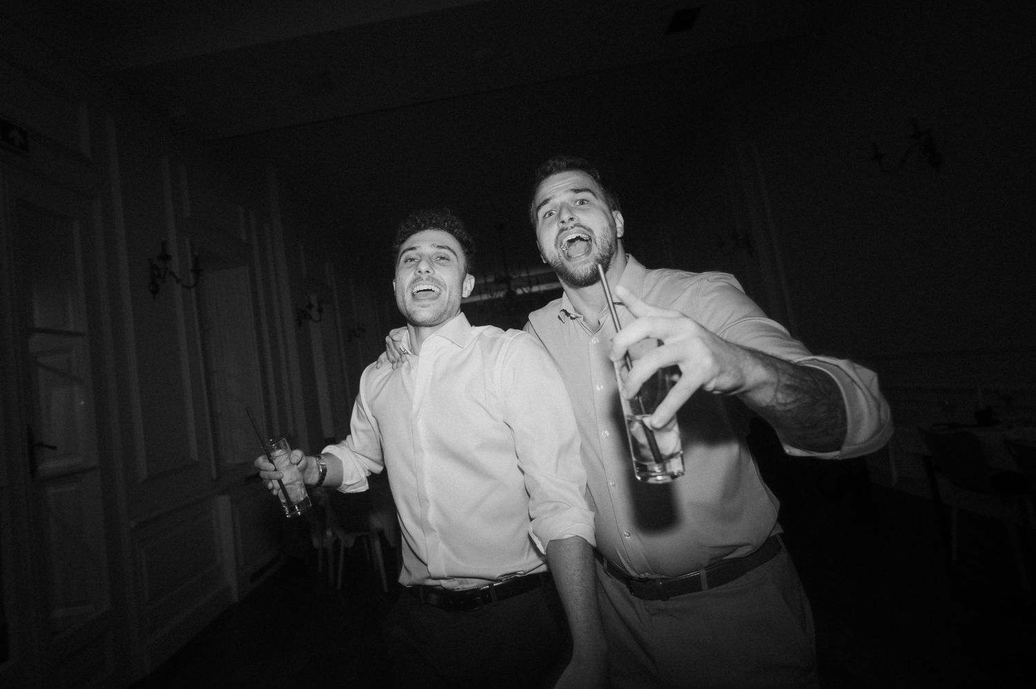 Two men laugh and celebrate with beer bottle at lively wedding reception in black and white.