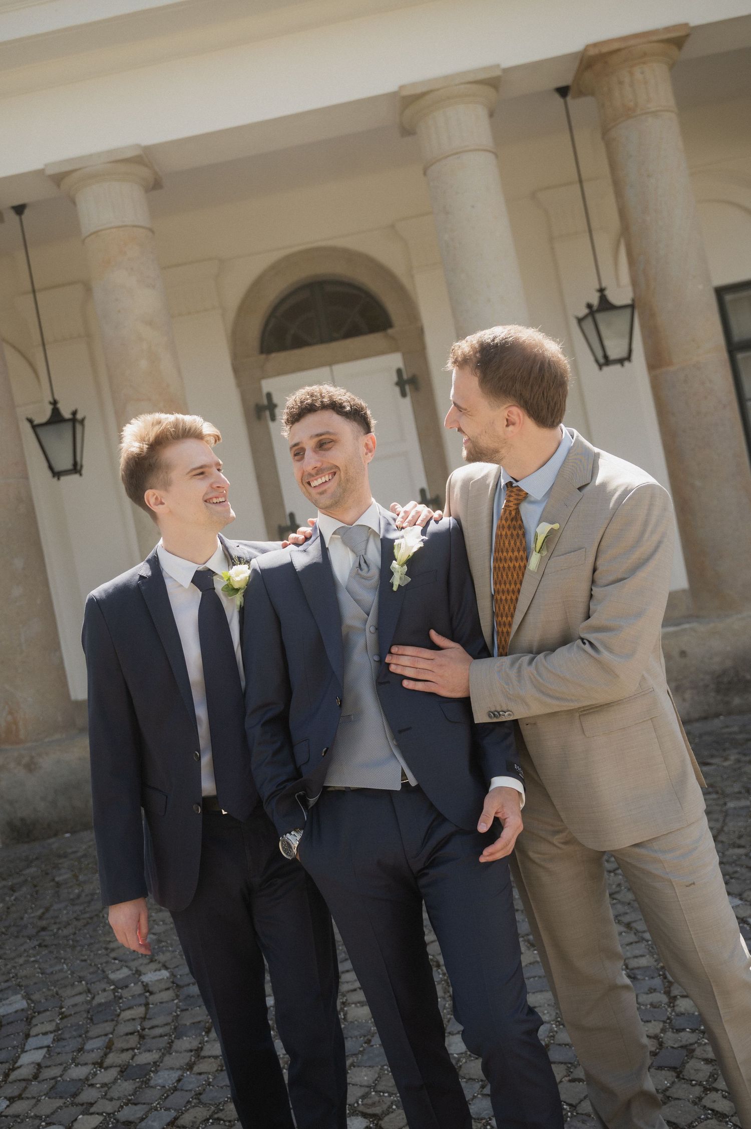 Three groomsmen in suits laughing and celebrating together outside a classical building on a wedding day.